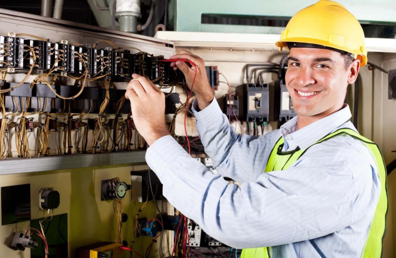 Engineer 桌布 壁紙 — 一位面帶微笑的電工，戴著黃色頭盔和綠色背心，正在仔細測試複雜電箱中的電線。