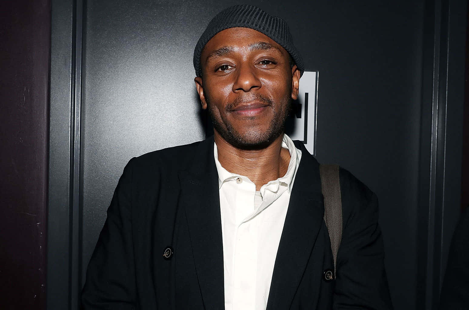 Yasiin Bey 桌布 壁紙 — 一位面帶溫暖笑容的男子，在深色背景前穿著黑色冷帽和西裝外套，內搭白色襯衫。