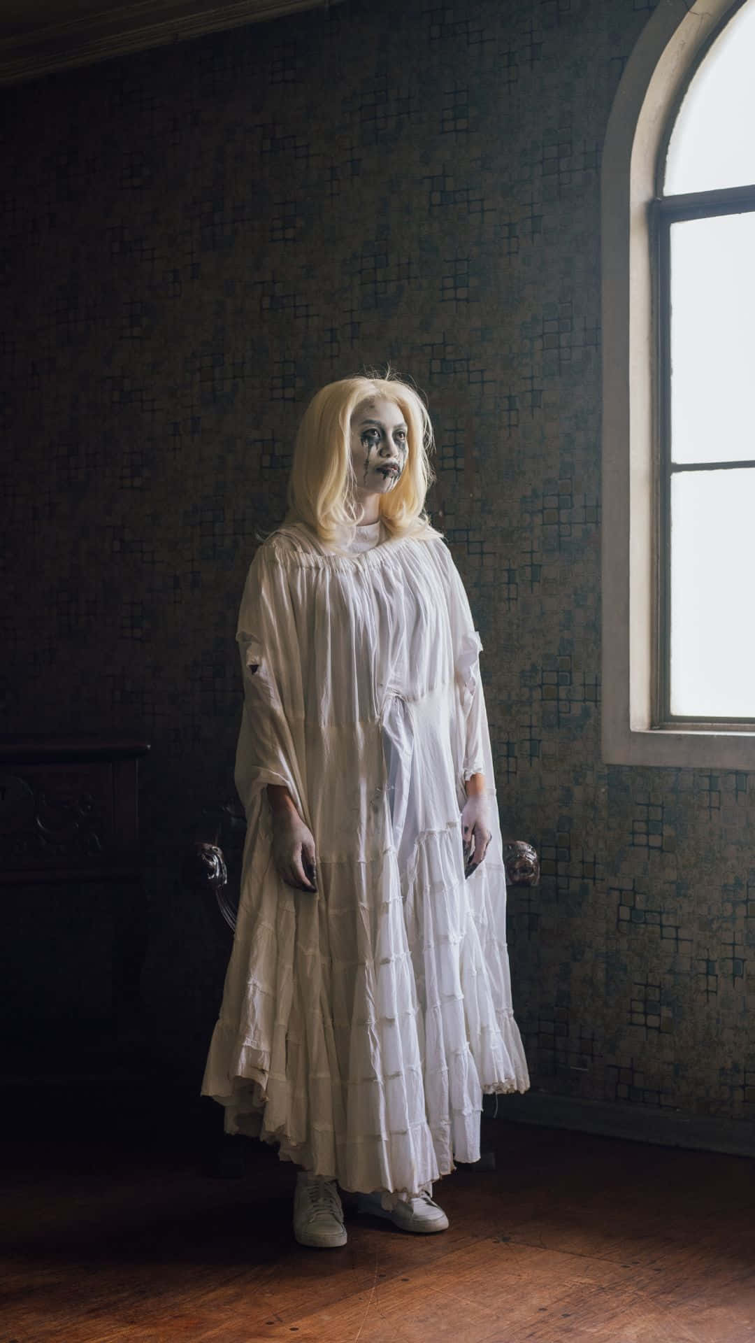Horror Costumes 桌布 壁紙 — 一位面色蒼白的女人穿著破爛的白裙，詭異地站在一個光線昏暗、有圖案牆紙和拱形窗戶的房間裡。