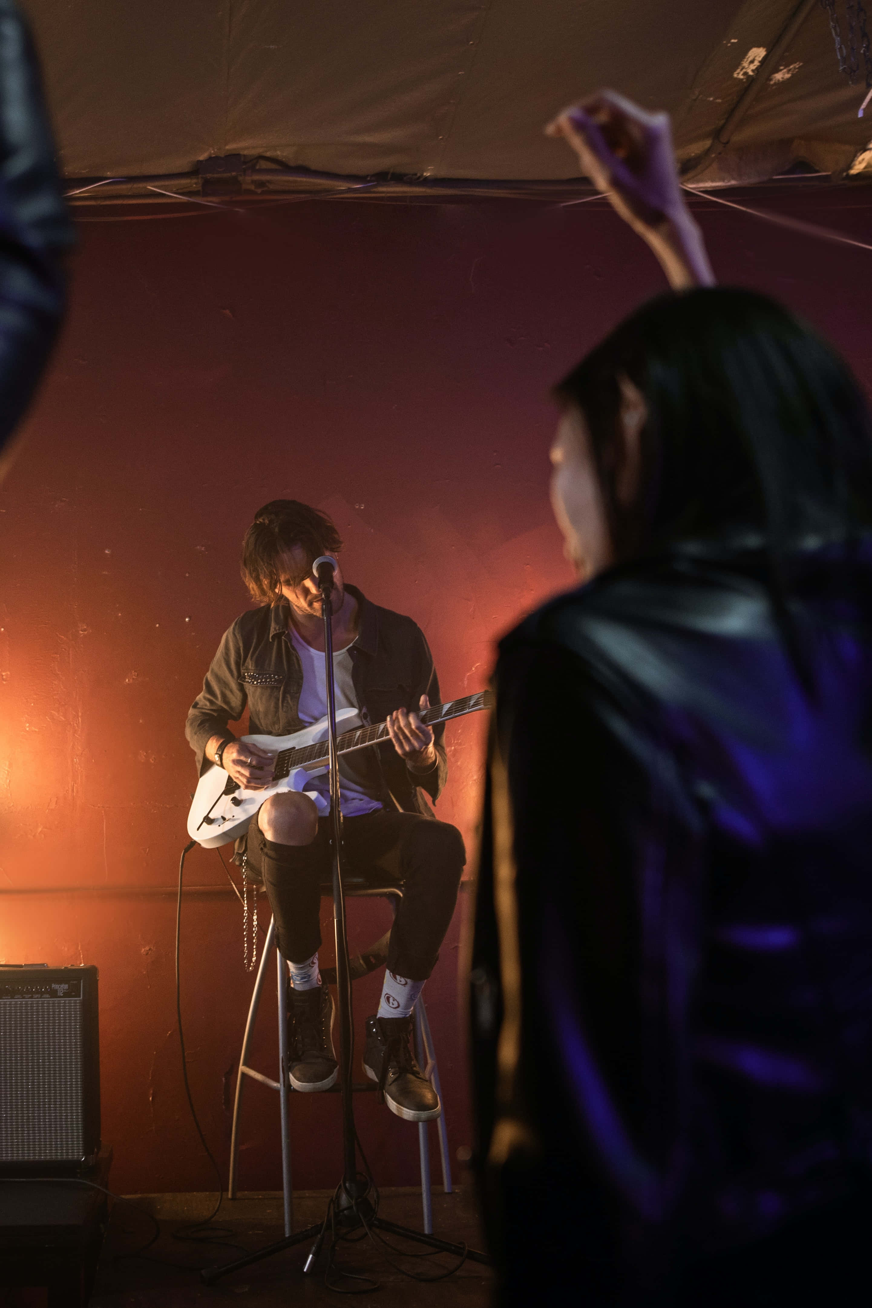 Singer 桌布 壁紙 — 一位音樂家在溫暖的舞台燈光下坐在凳子上彈奏電結他，而一位觀眾在紅牆前舉起手。