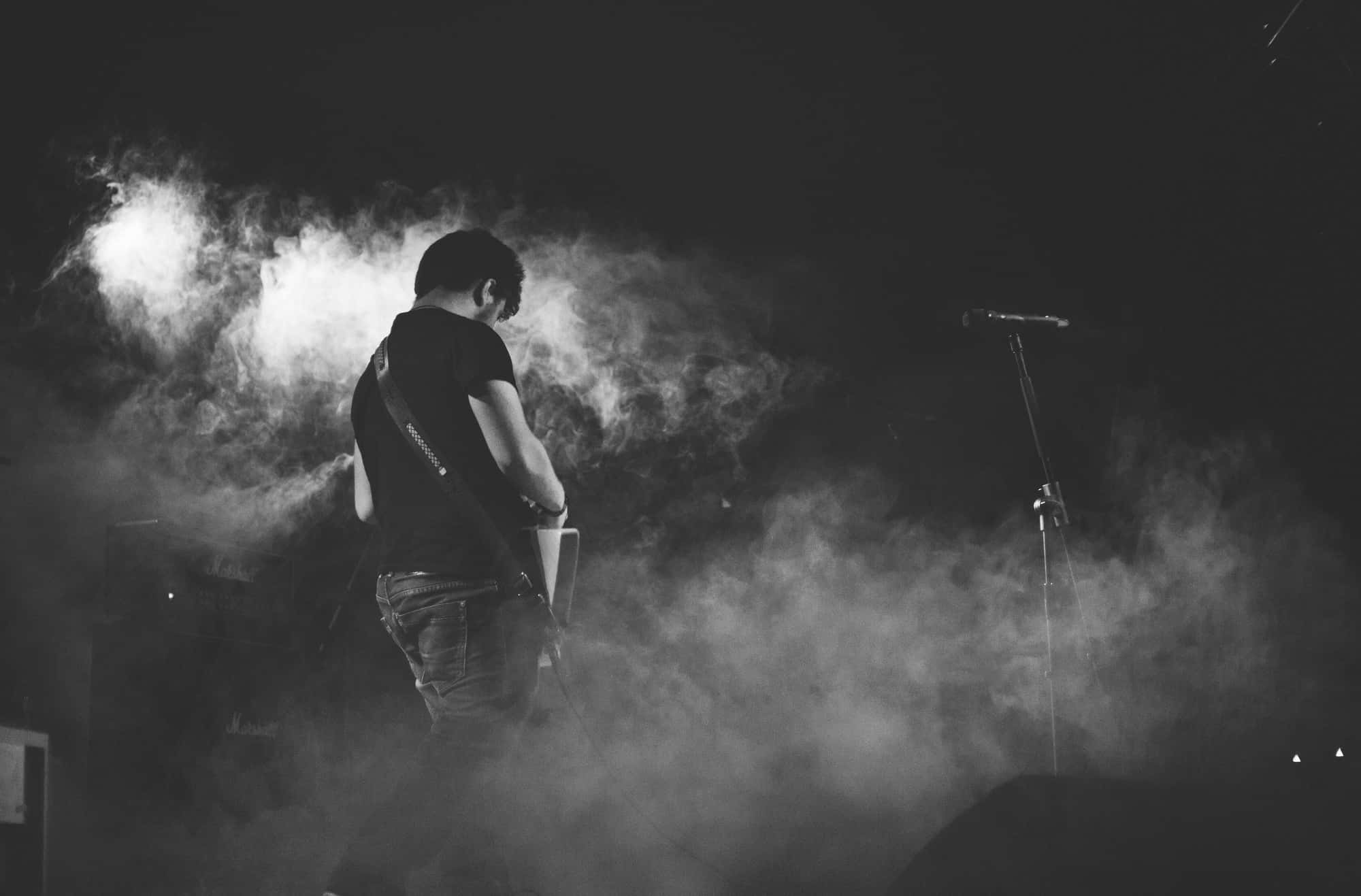 Singer 桌布 壁紙 — 一位音樂家在煙霧彌漫的舞台上，在戲劇性燈光下彈結他的黑白演唱會照片。