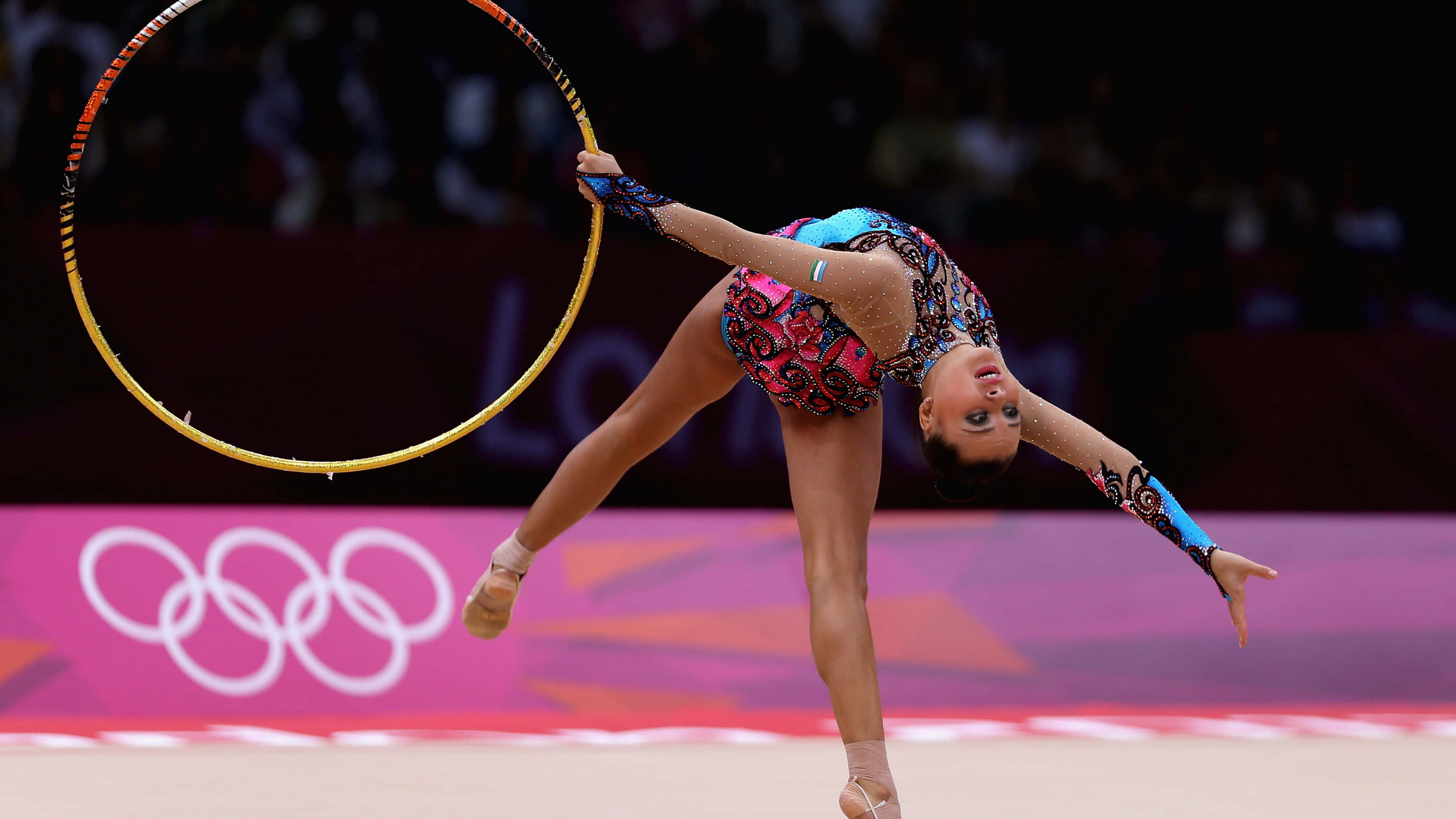 Gymnastics 桌布 壁紙 — 一位韻律體操運動員在燈光明亮的場館中，單腳優雅地平衡，同時旋轉一個金色圈環。