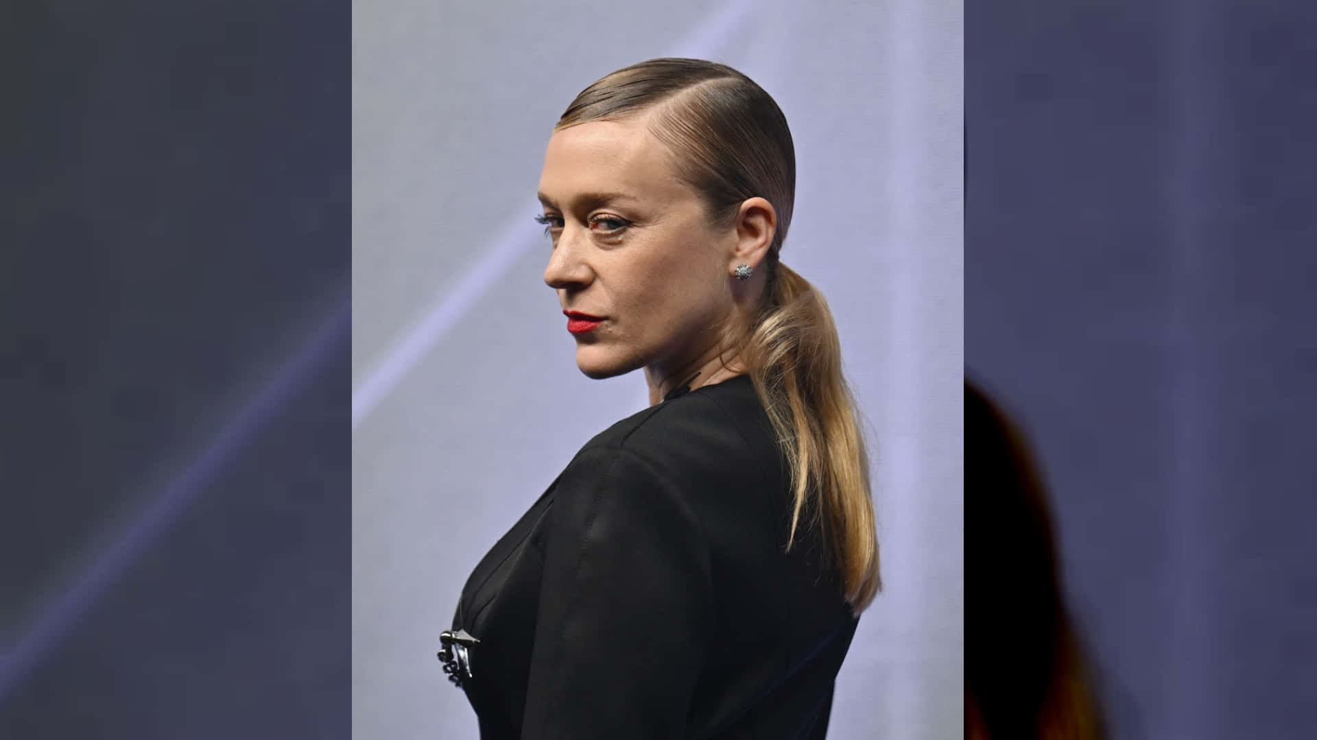 Chloë Sevigny 桌布 壁紙 — 一位頭髮向後梳、紅唇的女性，在柔和的藍色背景前側臉轉頭。