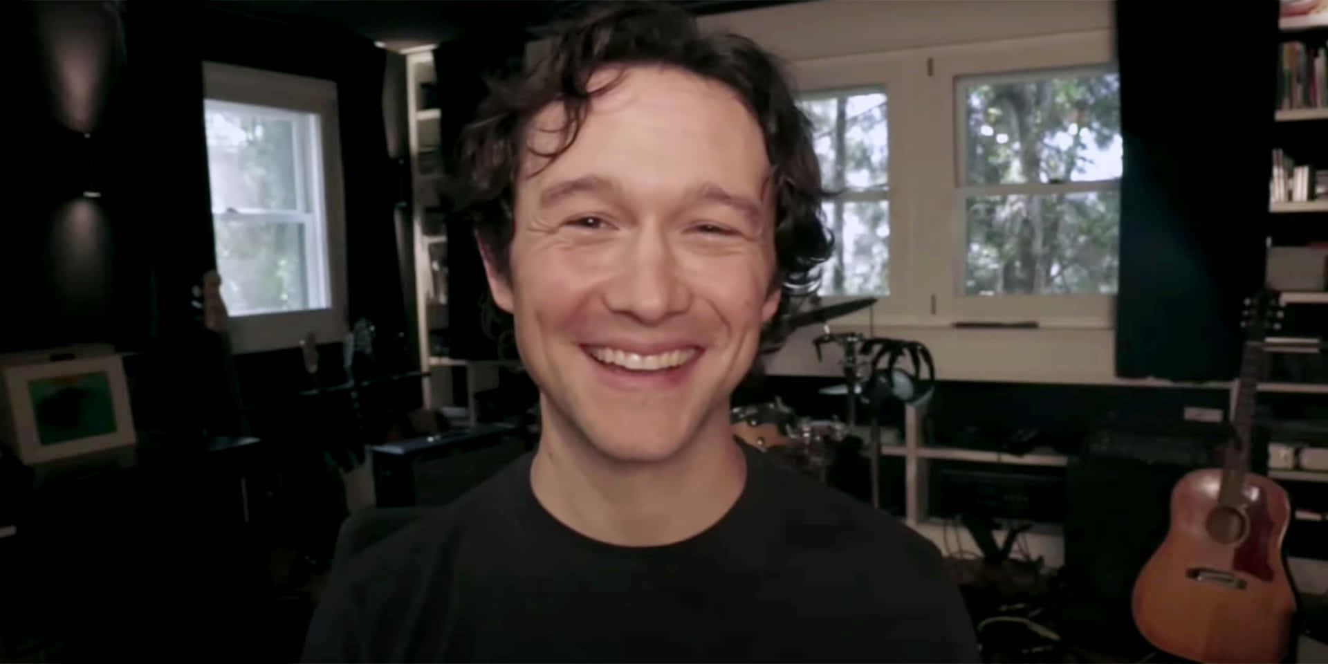 joseph gordon-levitt 桌布 壁紙 — 一位頭髮捲曲的開朗男子在光線昏暗、充滿樂器和窗戶的音樂錄音室裡溫暖地微笑。