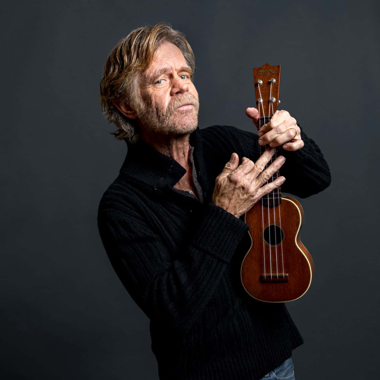 william h. macy 桌布 壁紙 — 一位頭髮花白、留鬍鬚的男子，在深灰色攝影棚背景前拿著一把木製烏克麗麗。