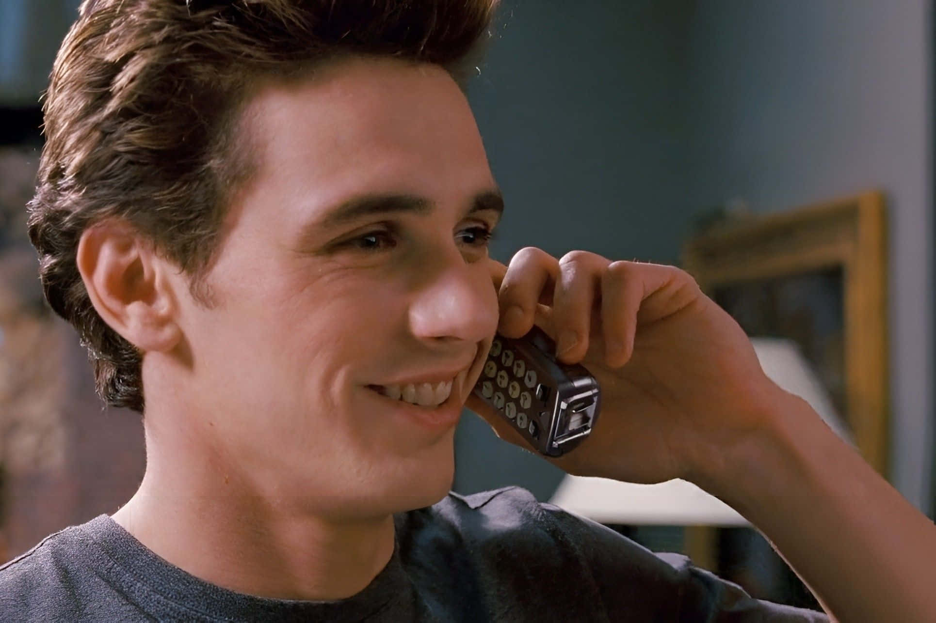 Harry Osborn 桌布 壁紙 — 一位頭髮蓬鬆的年輕男子在室內將舊款摺疊手機貼在耳邊，露出溫暖的笑容。