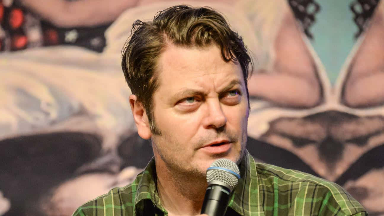 nick offerman 桌布 壁紙 — 一位頭髮造型時尚的男子，穿著綠色格仔襯衫，在模糊的壁畫背景下對著麥克風說話。