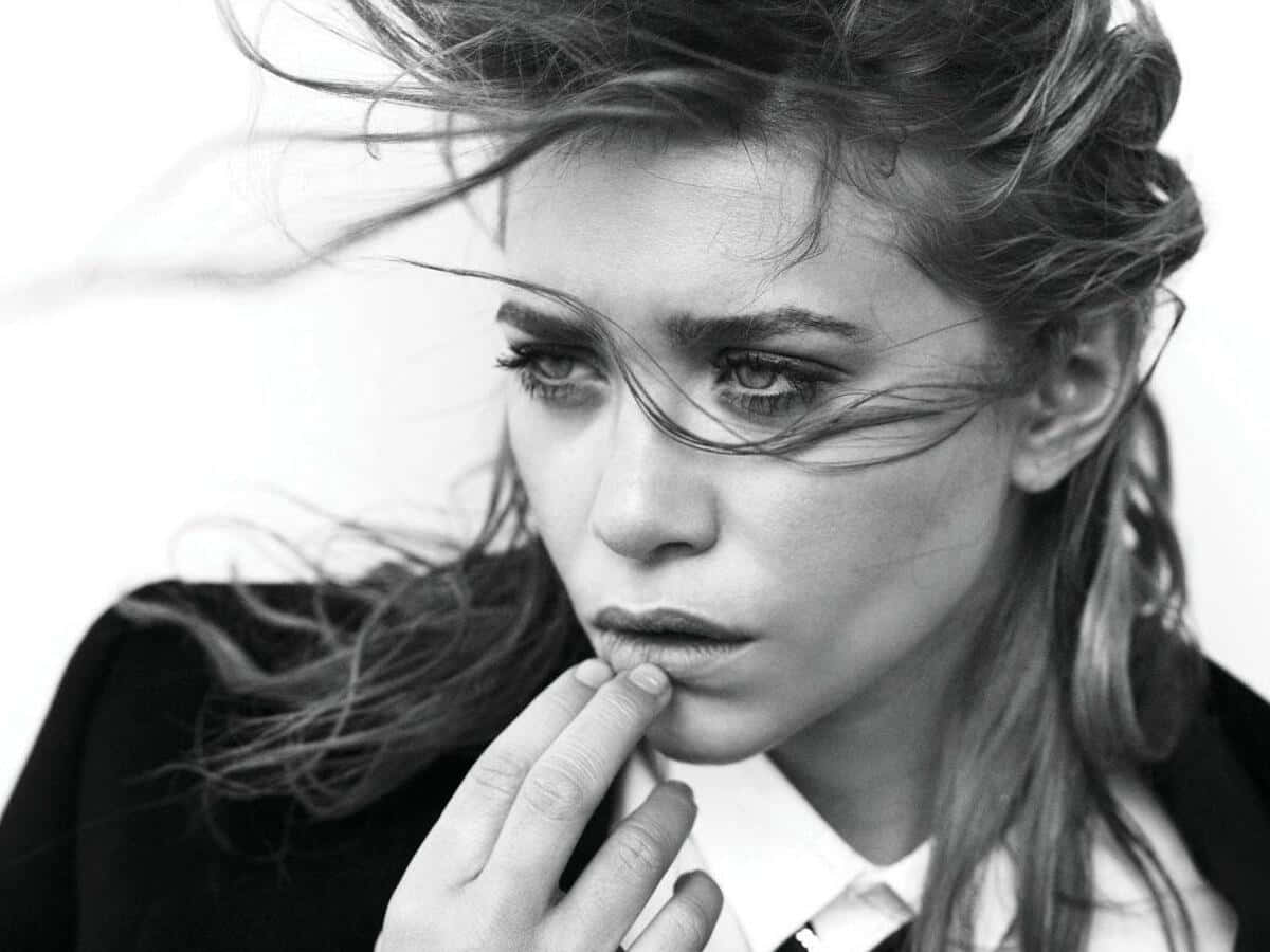 Mary Kate Olsen 桌布 壁紙 — 一位頭髮隨風飄揚的女性，若有所思地凝視著，同時觸摸嘴唇的黑白肖像。