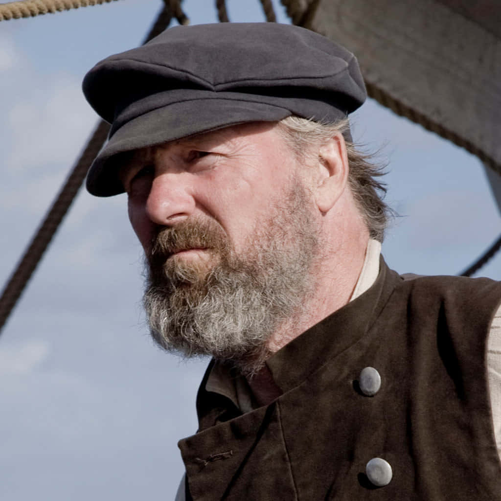 William Hurt 桌布 壁紙 — 一位飽經風霜的水手，留著花白的鬍鬚，戴著帽子，在船隻索具和藍天的背景下若有所思地凝視著。