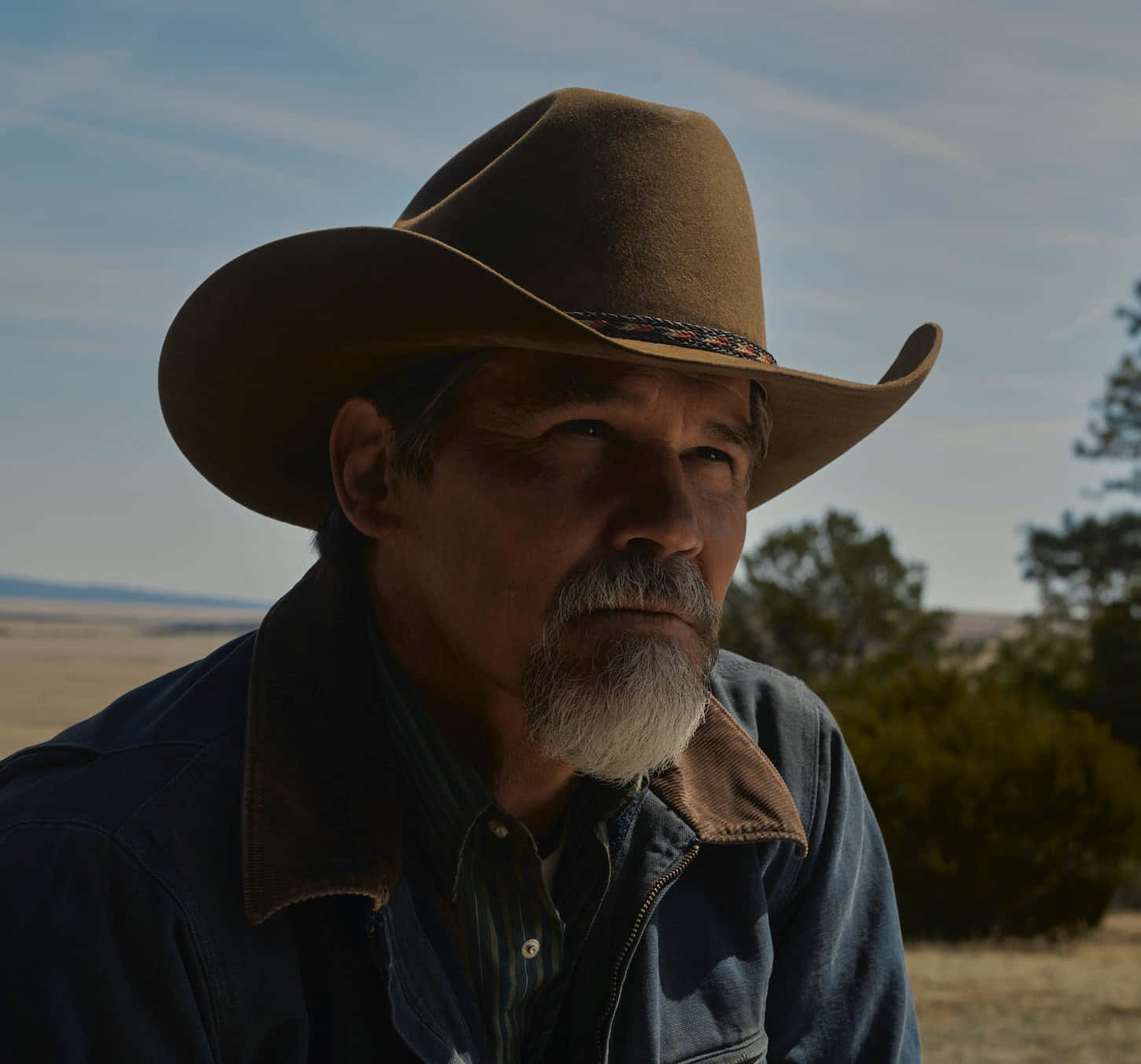 Josh Brolin 桌布 壁紙 — 一位飽經風霜的牛仔，留著白鬍子，戴著棕色帽子，在晴朗的藍天下，對著廣闊的沙漠背景若有所思地凝視。