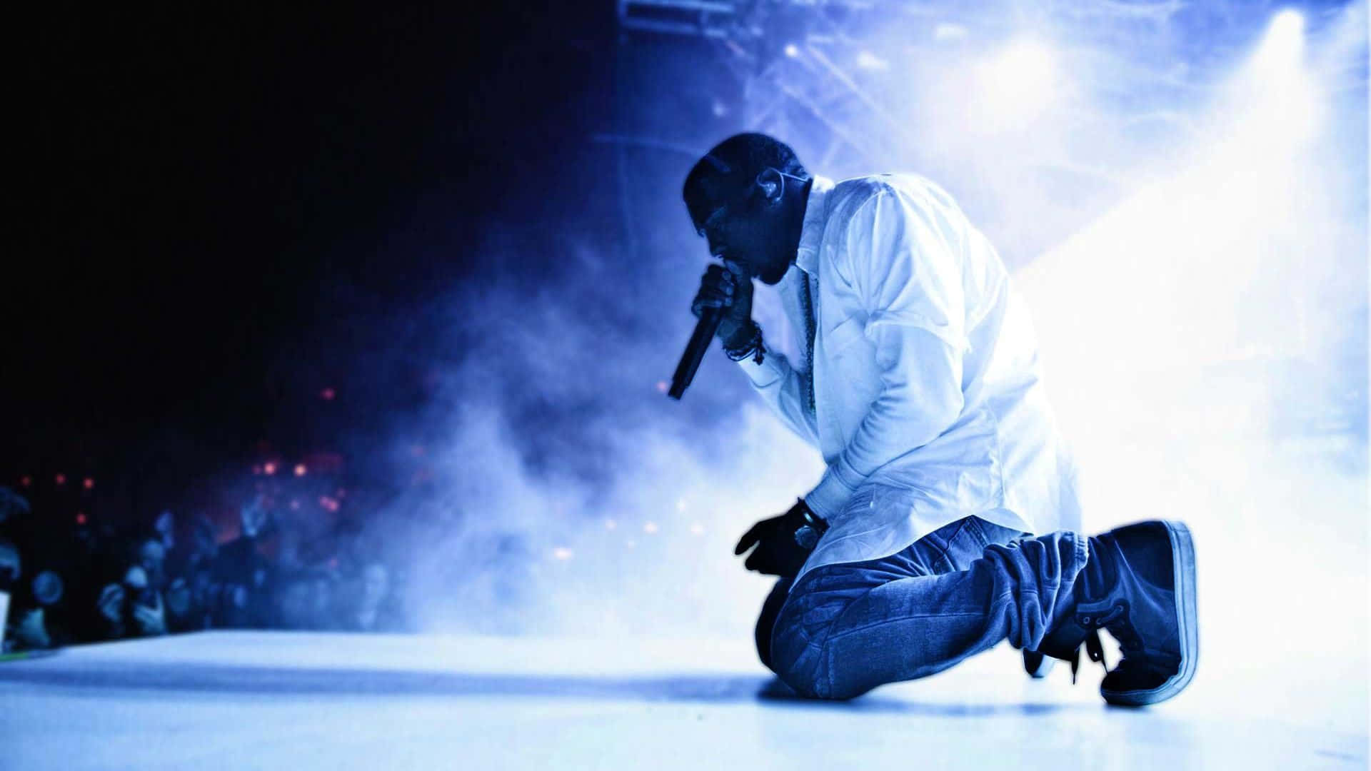 Kanye 桌布 壁紙 — 一位饒舌歌手跪在舞台上，沐浴在藍光中，煙霧在他周圍旋轉，他對著麥克風表演。