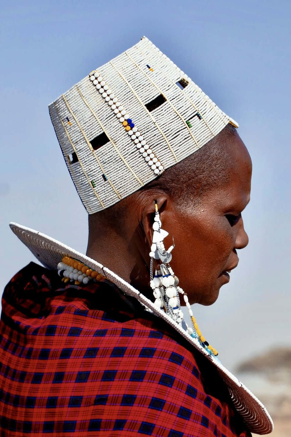 African Culture 桌布 壁紙 — 一位馬賽族女性側面照，戴著串珠白色帽子和紅色格紋披肩，背景是清澈的藍天。