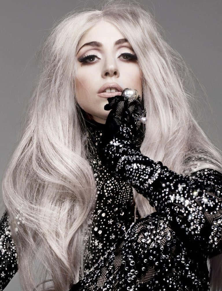 Lady Gaga 桌布 壁紙 — 一位魅力四射的女性，擁有長銀色頭髮和戲劇性妝容，穿著閃亮的黑色服裝和珍珠裝飾手套，在灰色背景前擺姿勢。
