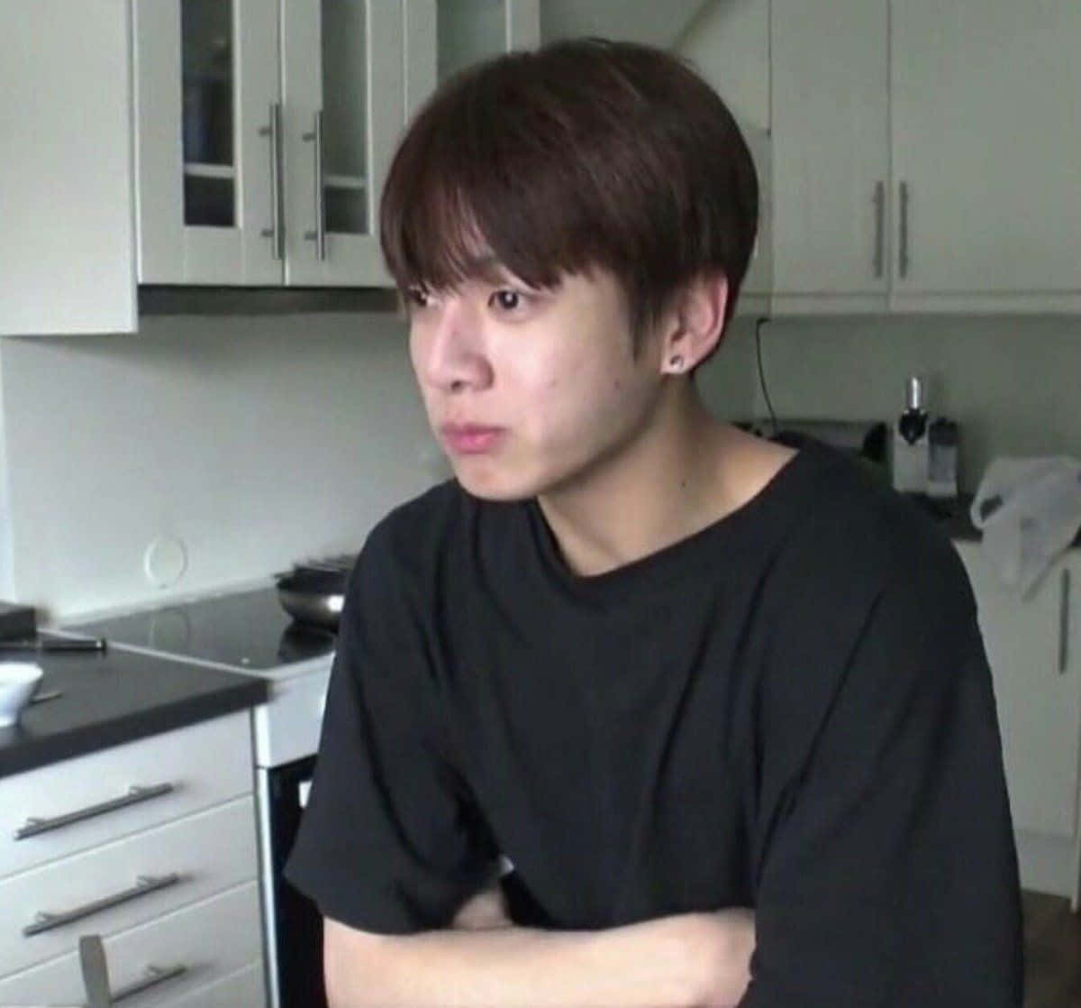 Bts Meme 桌布 壁紙 — 一位黑髮、身穿黑色T恤的年輕男子坐在一個有白色櫥櫃和黑色枱面的現代廚房裏。