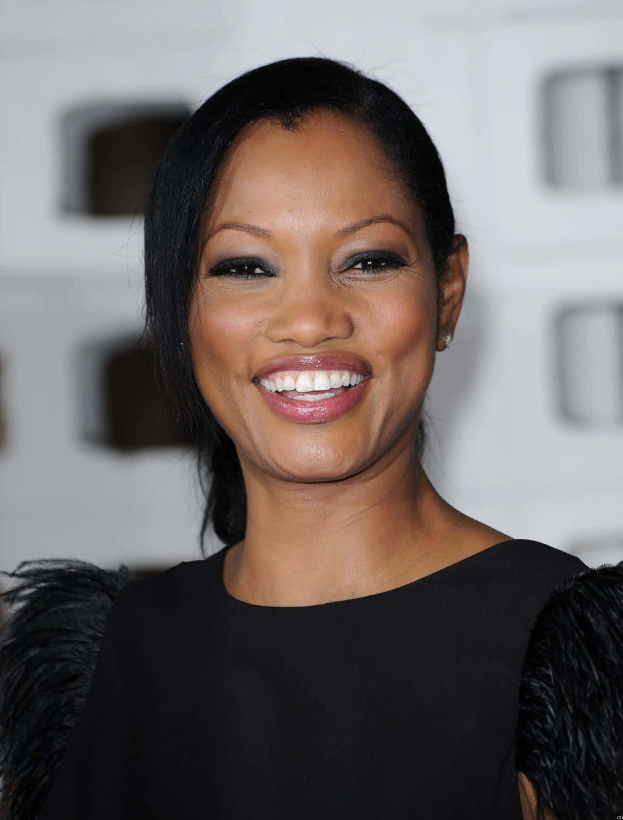 Garcelle Beauvais 桌布 壁紙 — 一位黑髮亮麗、笑容燦爛的女性，身穿黑色羽毛連衣裙，背景是模糊的幾何圖案。