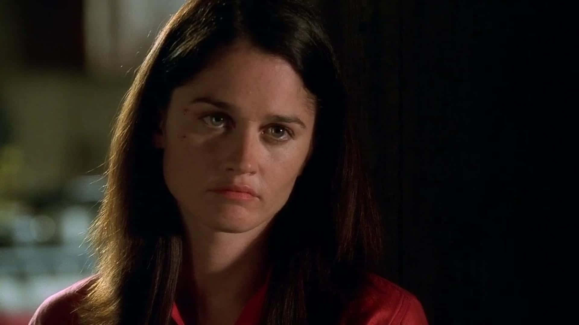 Robin Tunney 桌布 壁紙 — 一位黑髮女子身穿紅色襯衫，在光線昏暗的房間裡專注地凝視，背景柔和模糊。