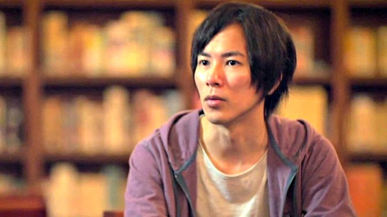 Isayama Hajime 桌布 壁紙 — 一位黑髮年輕男子在溫暖燈光的圖書館中，被書架環繞，若有所思地坐著。