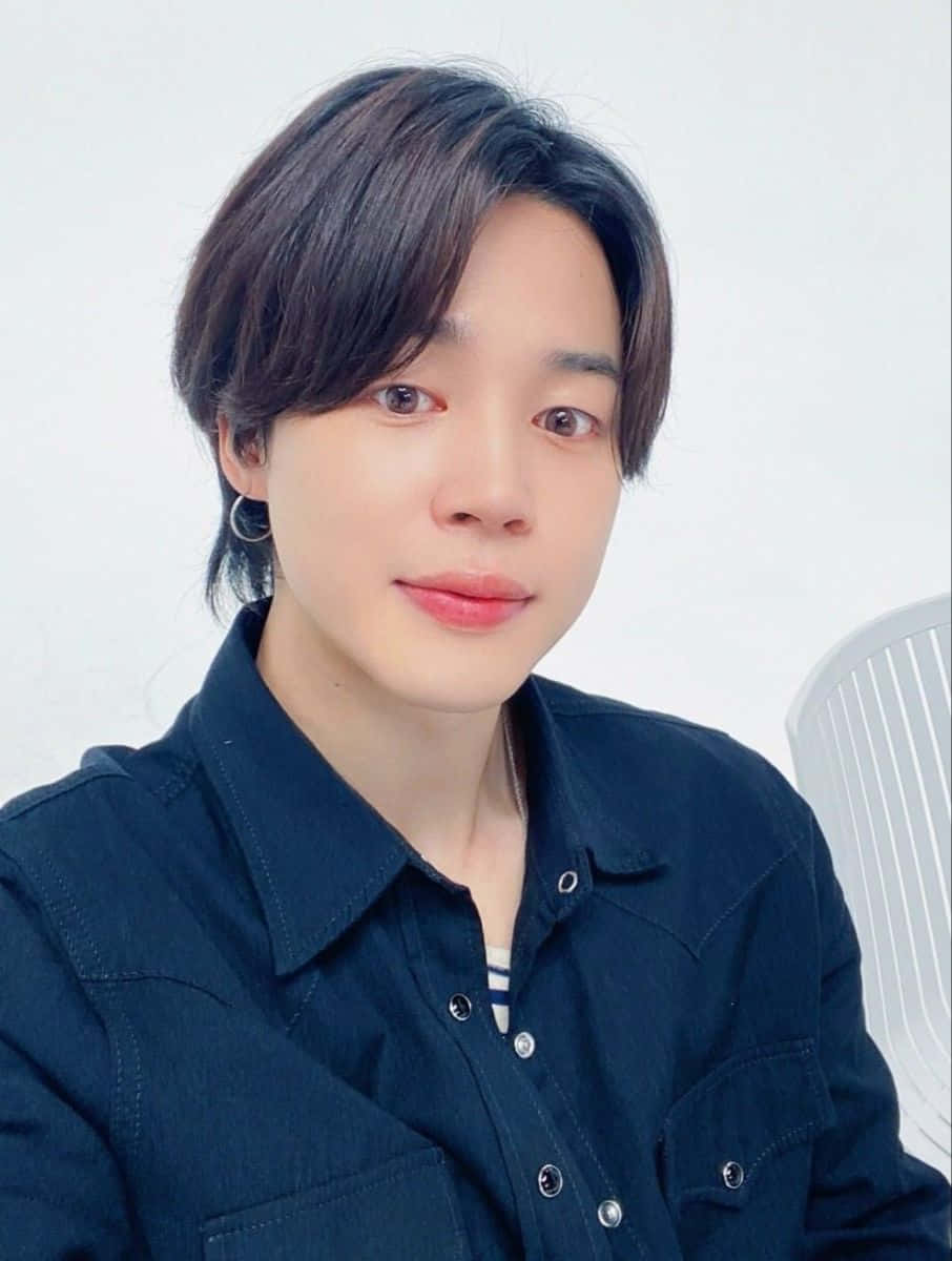 Bts Endorsements 桌布 壁紙 — 一位黑髮戴銀色耳環的年輕男子，穿著深色牛仔襯衫，在純白背景前溫柔微笑。