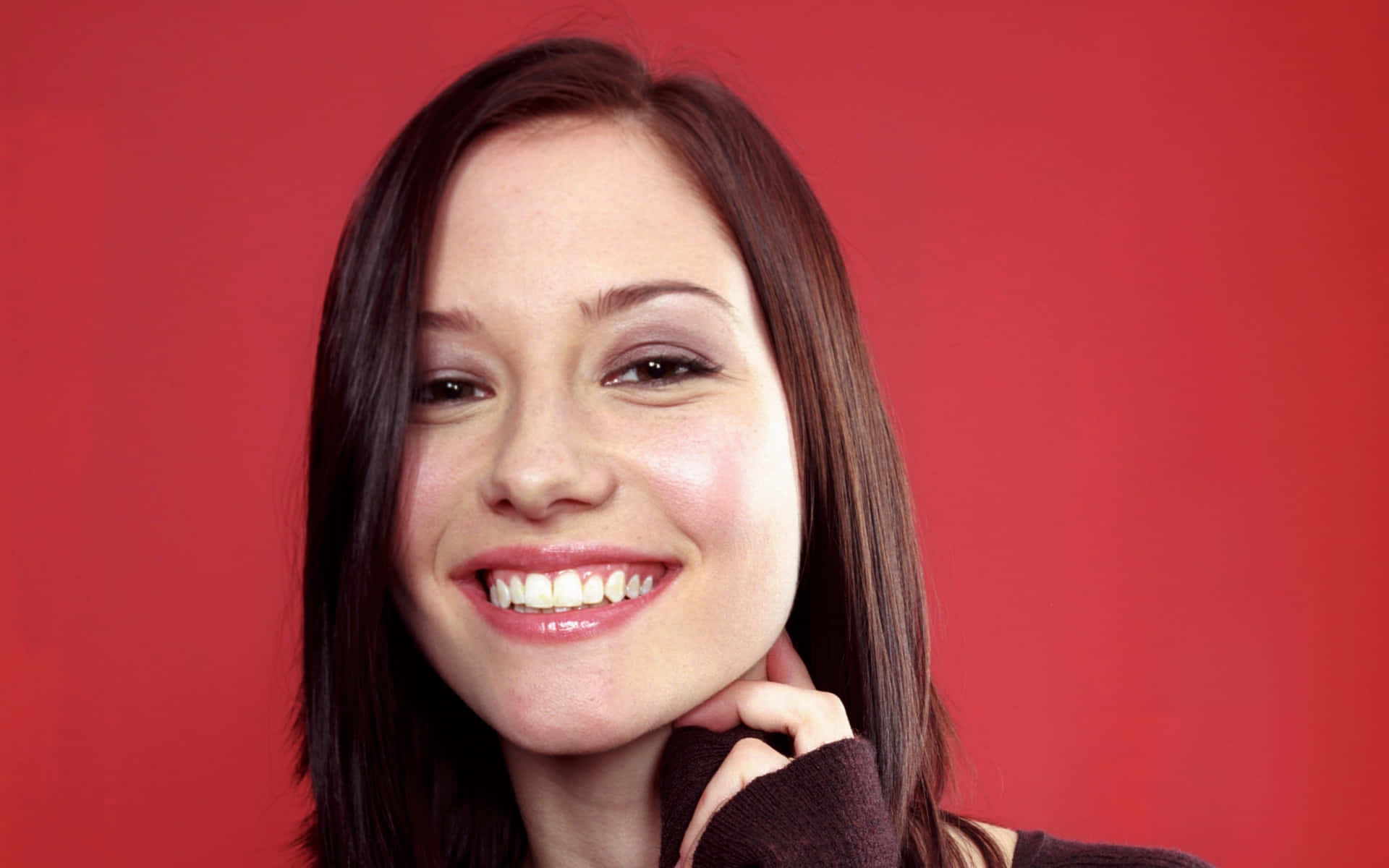 Chyler Leigh 桌布 壁紙 — 一位黑髮的開朗女性在純紅色背景前溫暖微笑，她的手輕輕觸碰脖子。