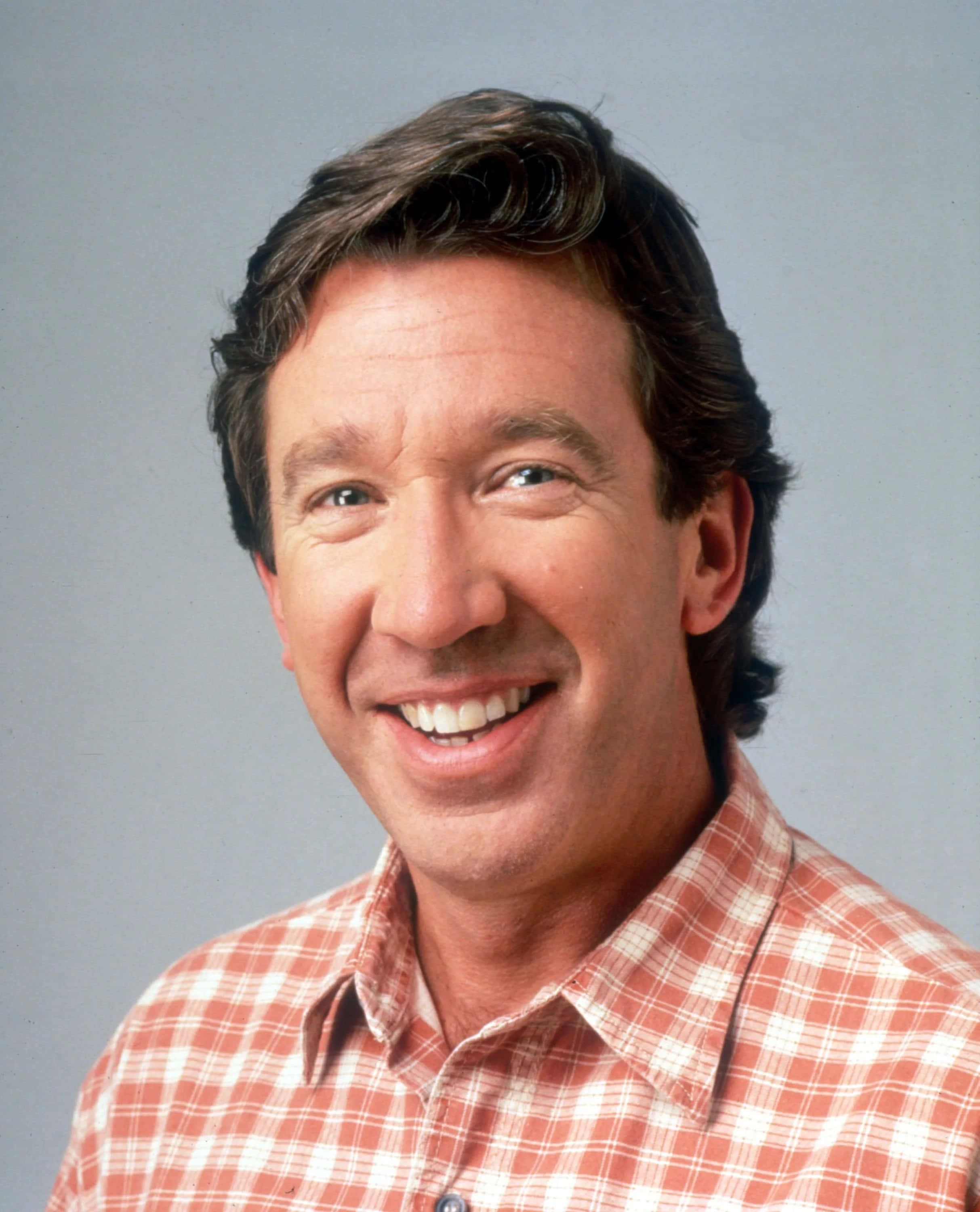 Tim Allen 桌布 壁紙 — 一位黑髮的開朗男子在純灰色攝影棚背景前，穿著紅白格紋襯衫，露出燦爛的笑容。