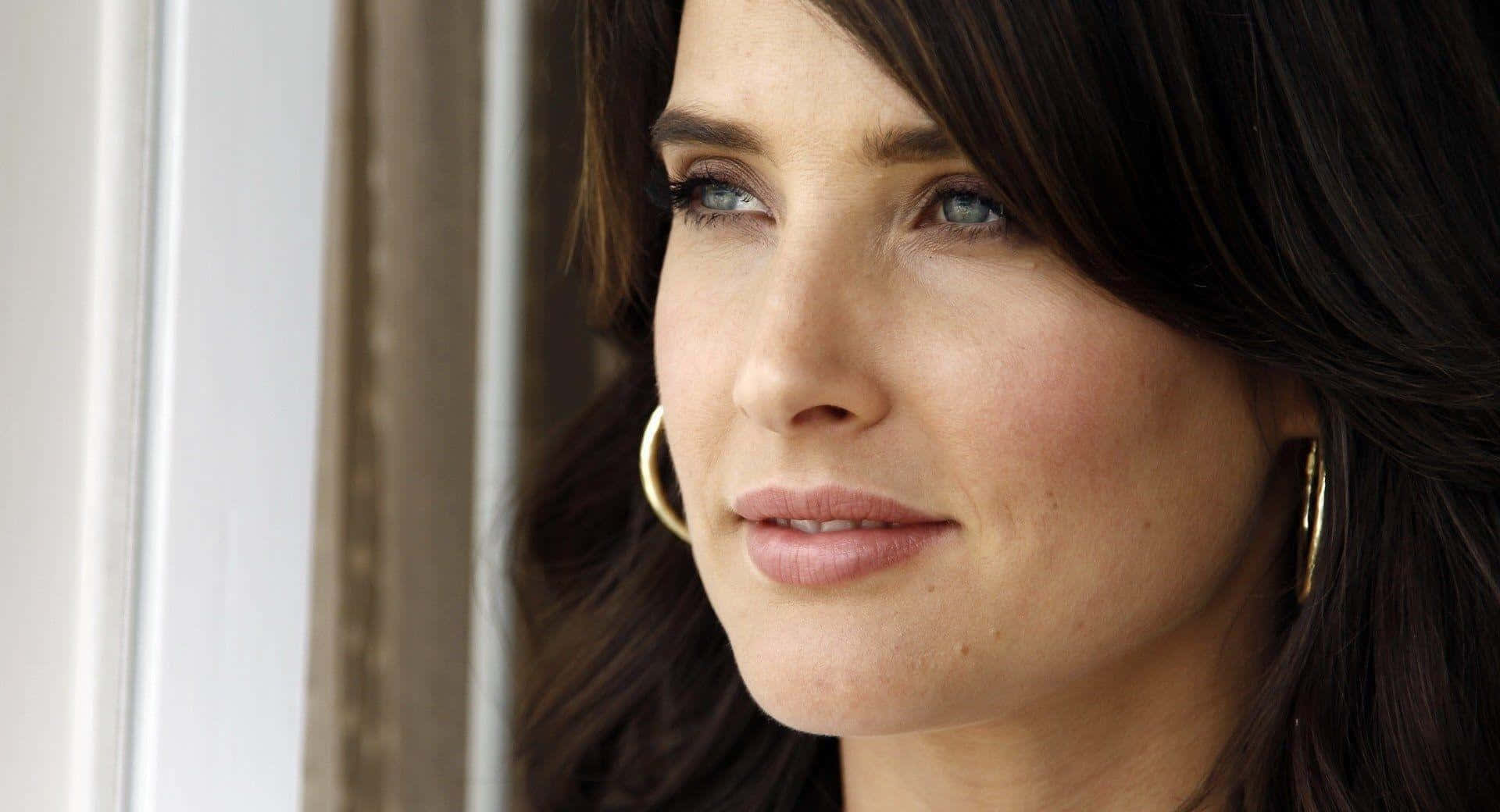 Cobie Smulders 桌布 壁紙 — 一位黑髮藍眼女士戴著金色圓環耳環，若有所思地凝視窗外的特寫肖像。