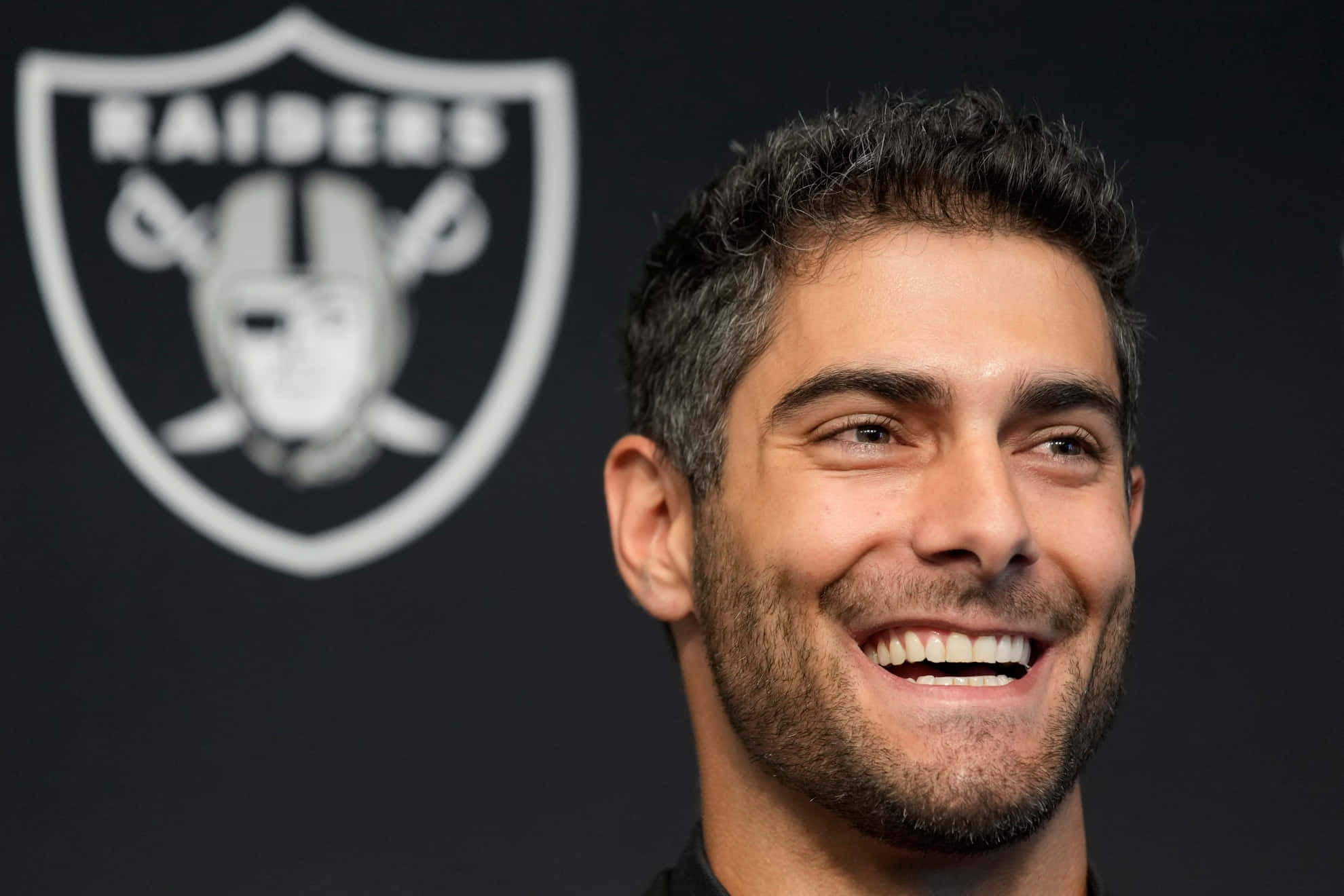 Jimmy Garoppolo 桌布 壁紙 — 一位黑髮鬍鬚的男子微笑著站在黑色背景前，背景上有白色的奧克蘭突襲者隊標誌。