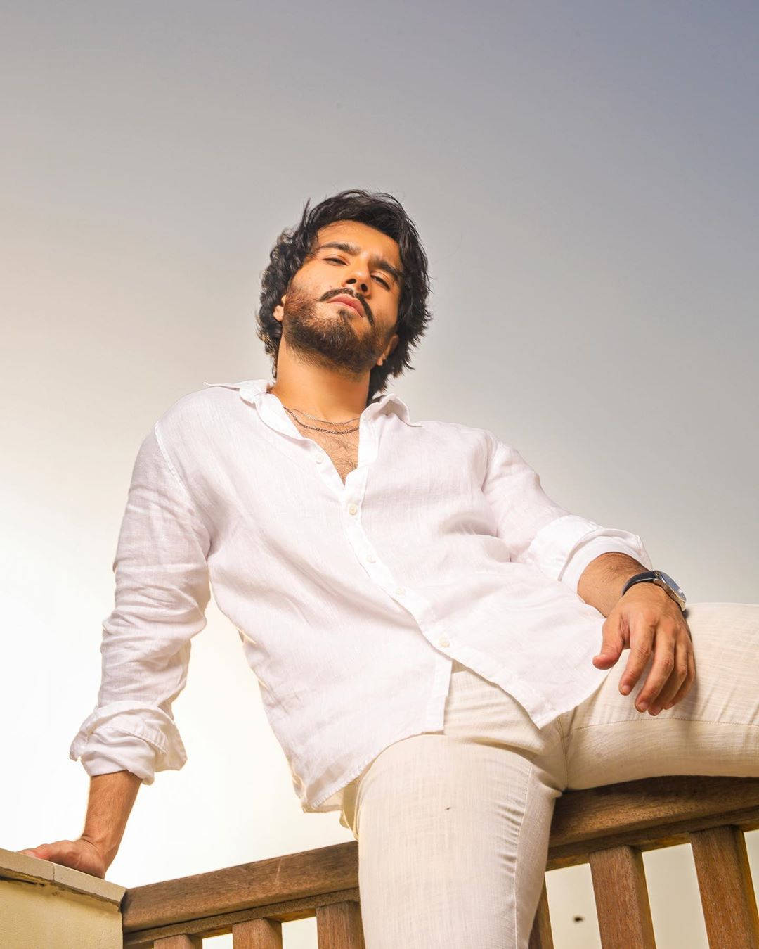 Feroz Khan 桌布 壁紙 — 一位黑髮鬍鬚的男子身穿白色亞麻衣服，靠著木欄杆，背景是柔和的天空。