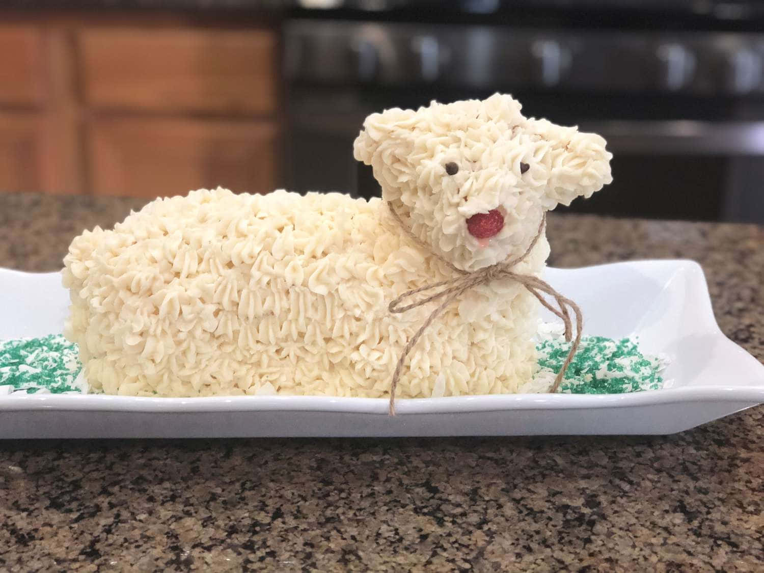 Lamb 桌布 壁紙 — 一個 fluffy 白色糖霜蛋糕，造型像羊，有紅色糖果鼻子和麻繩蝴蝶結，放在一個有綠色糖粒的白色碟子上。