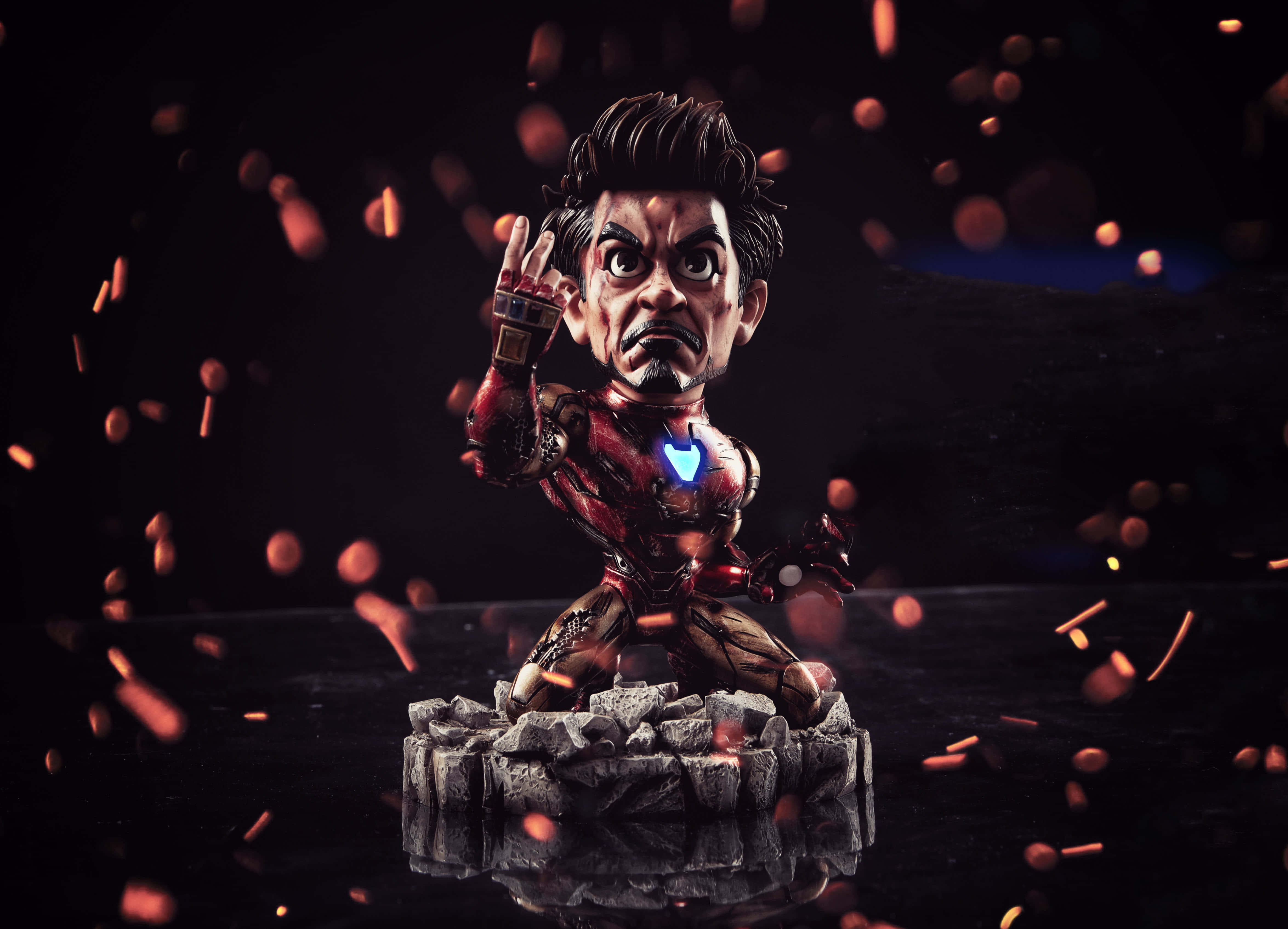 Iron Man Bobbleheads 桌布 壁紙 — 一個Q版風格的鐵甲奇俠人偶，在黑暗背景下，站在岩石底座上，周圍漂浮著火花，姿態挑釁。