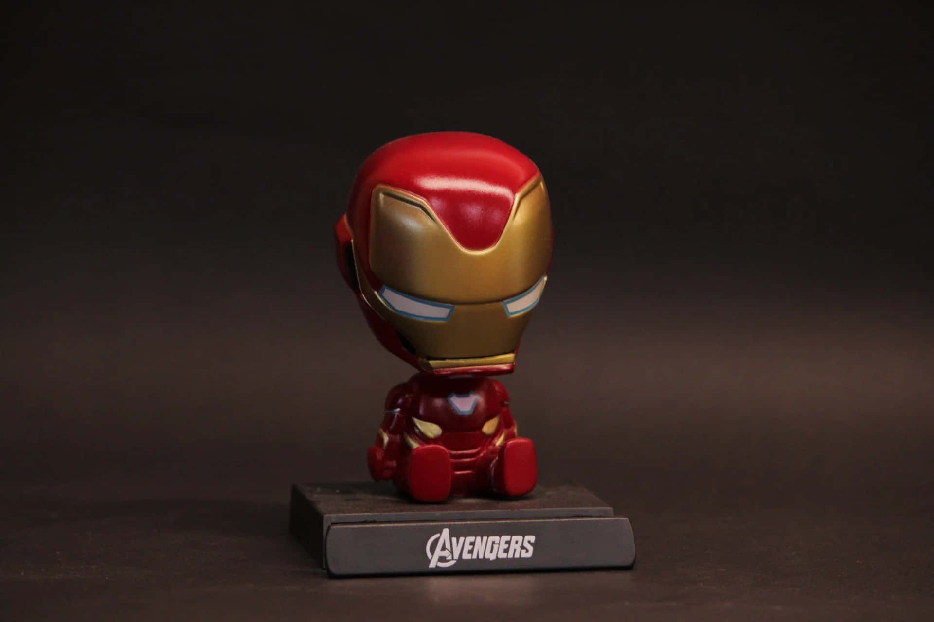 Iron Man Bobbleheads 桌布 壁紙 — 一個Q版風格的鐵甲奇俠人偶坐在黑色復仇者聯盟底座上，背景為深色攝影棚。