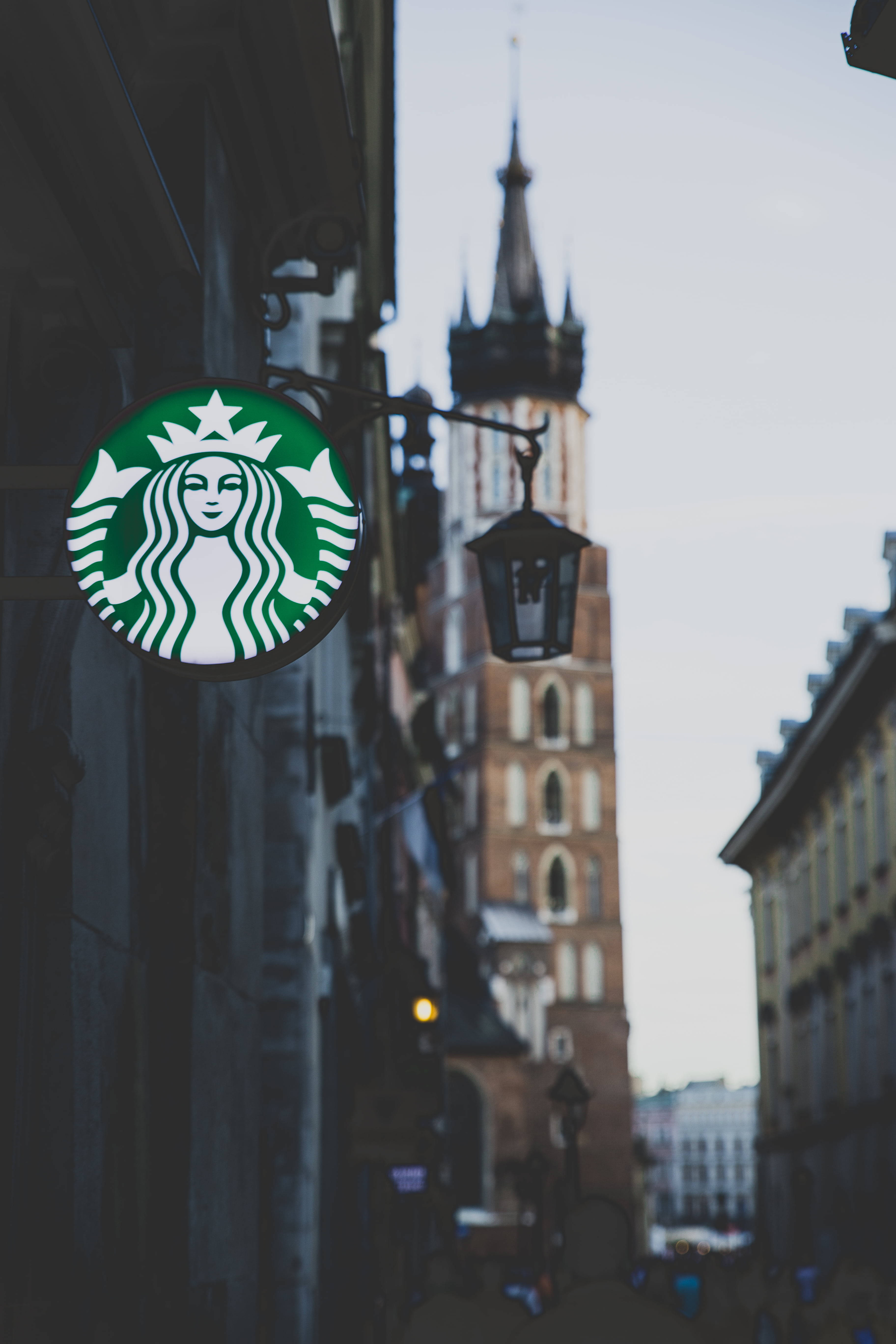 星巴克 桌布 壁紙 — 一個Starbucks標誌招牌掛在建築物上，背景模糊中可見一座歷史悠久的教堂塔樓。