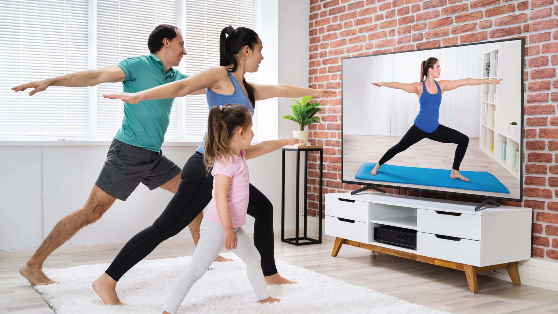 Home Entertainment 桌布 壁紙 — 一個三人家庭在明亮現代、有磚牆和白色地毯的客廳中，模仿電視上的瑜伽姿勢。