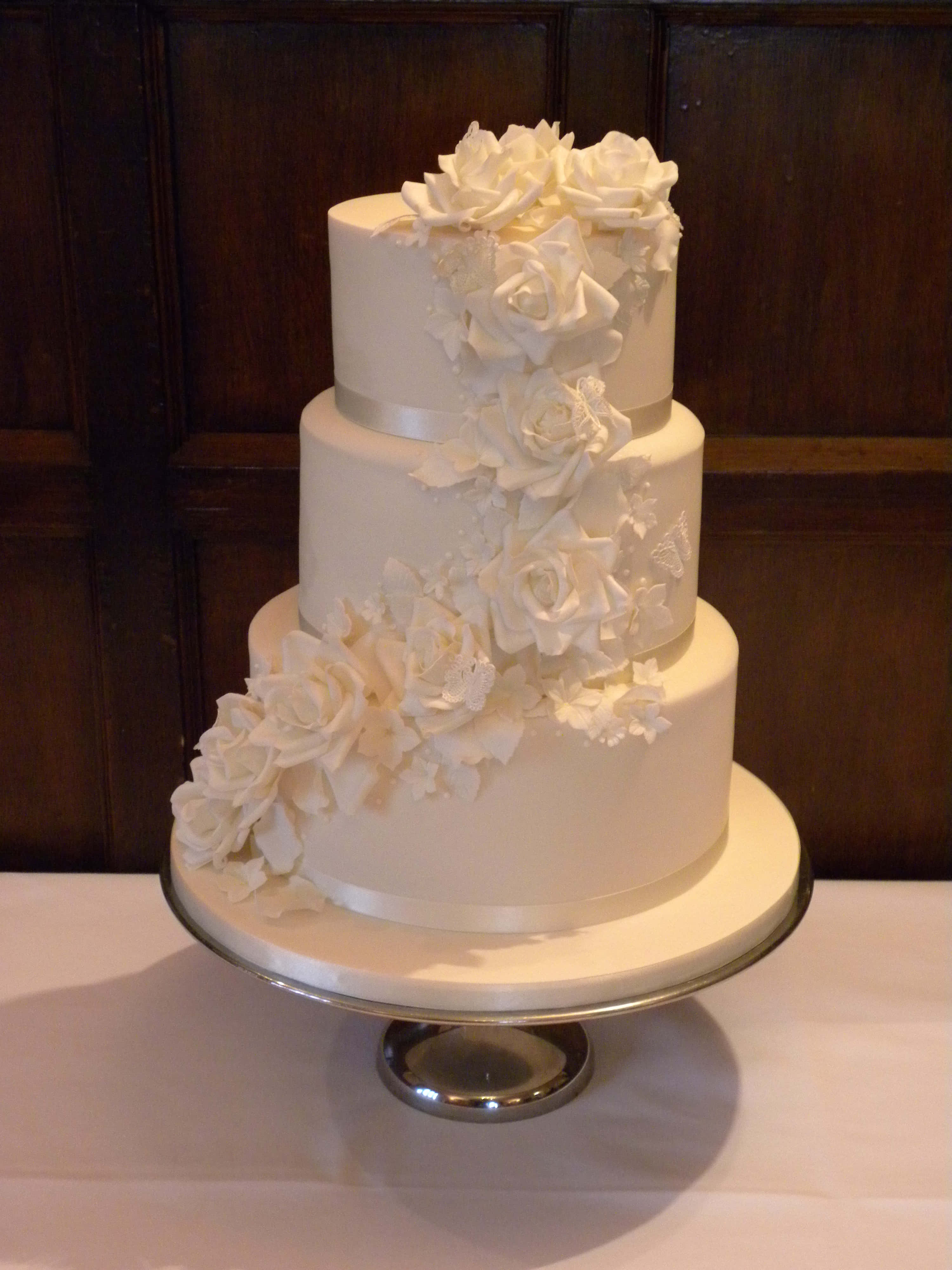 Wedding Cake 桌布 壁紙 — 一個三層白色結婚蛋糕，綴以層疊的糖玫瑰，優雅地放在銀色支架上，背景是深色木牆板。