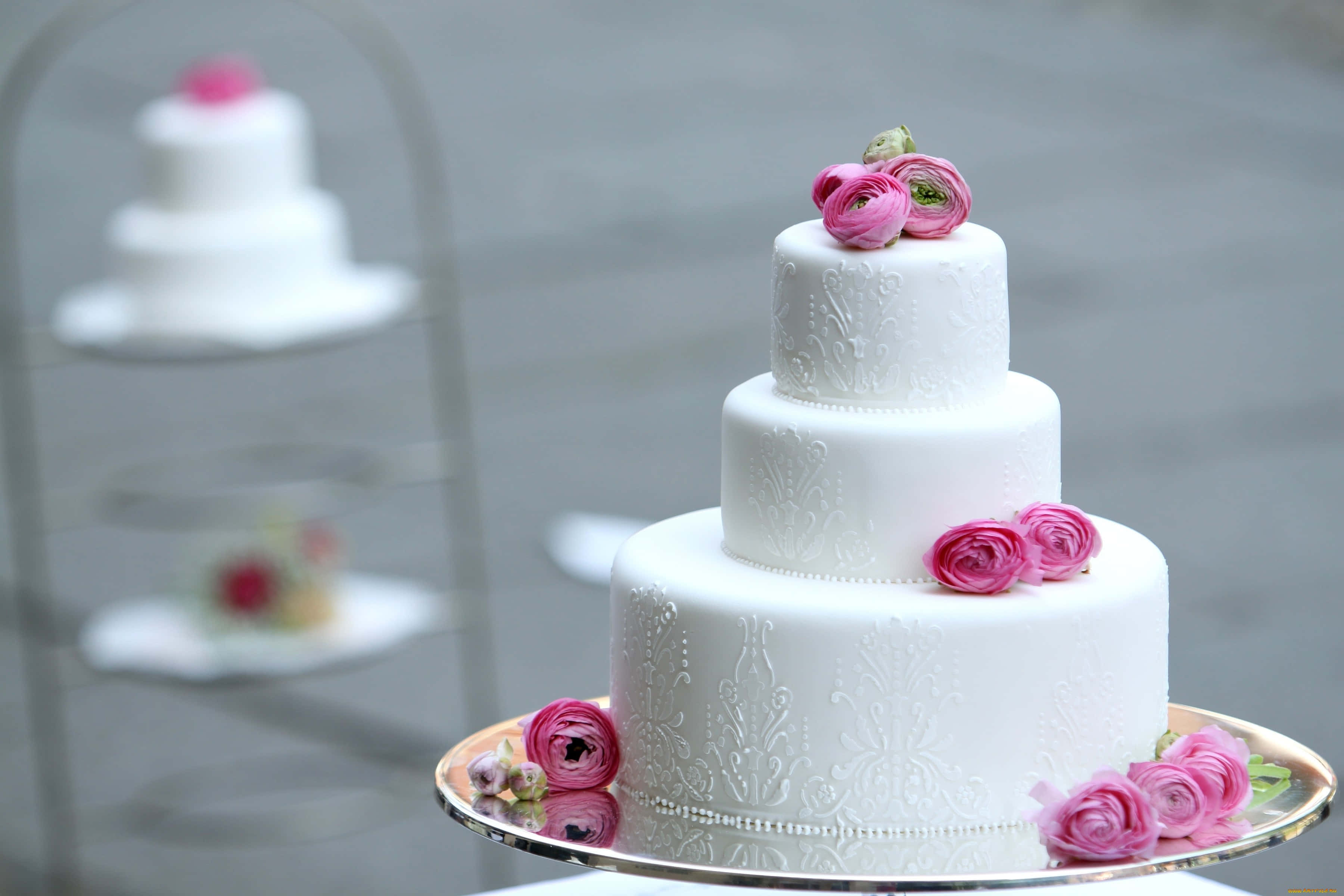 Floral Cake 桌布 壁紙 — 一個三層白色結婚蛋糕，飾以粉紅色毛茛花，優雅地放在銀盤上，背景有模糊的蛋糕架。