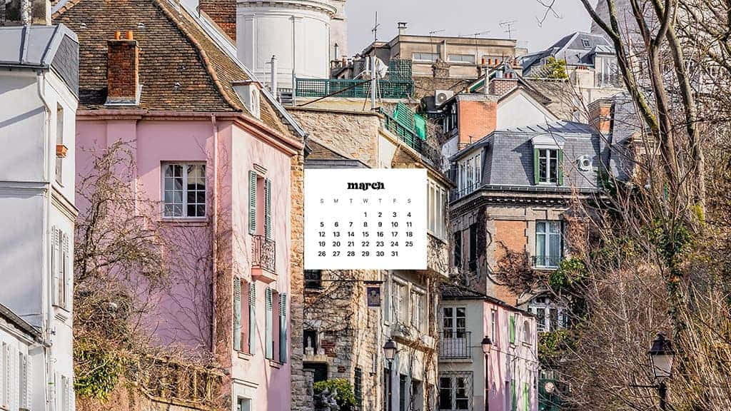 March Desktop 桌布 壁紙 — 一個三月日曆漂浮在柔和日光下、擁有瓦片屋頂和光禿樹木的粉彩歐洲房屋之間。