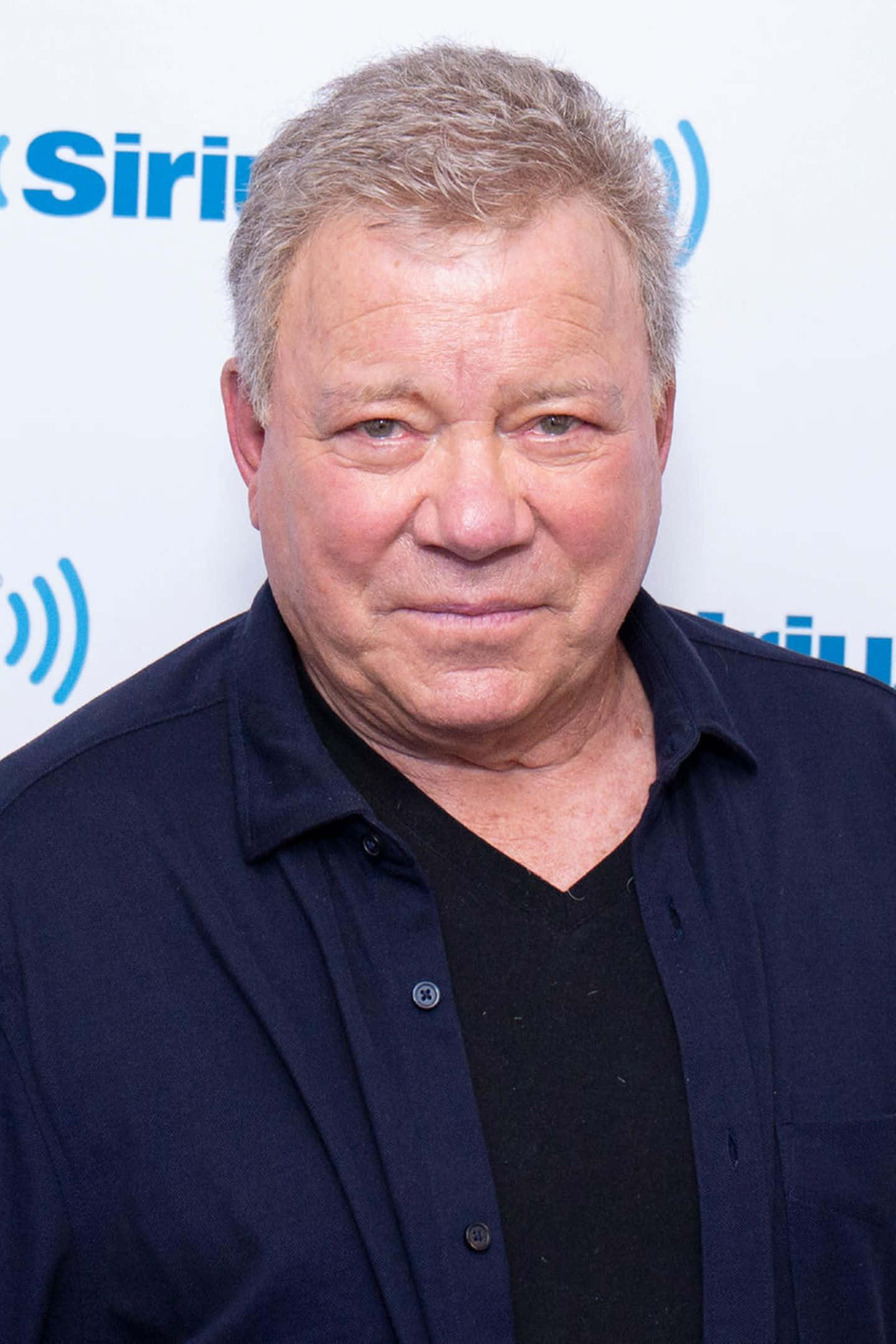 william shatner 桌布 壁紙 — 一個中年男人嘅特寫肖像，佢有灰白頭髮，喺SiriusXM活動入面著住一件深藍色鈕扣恤衫，入面襯黑色V領。