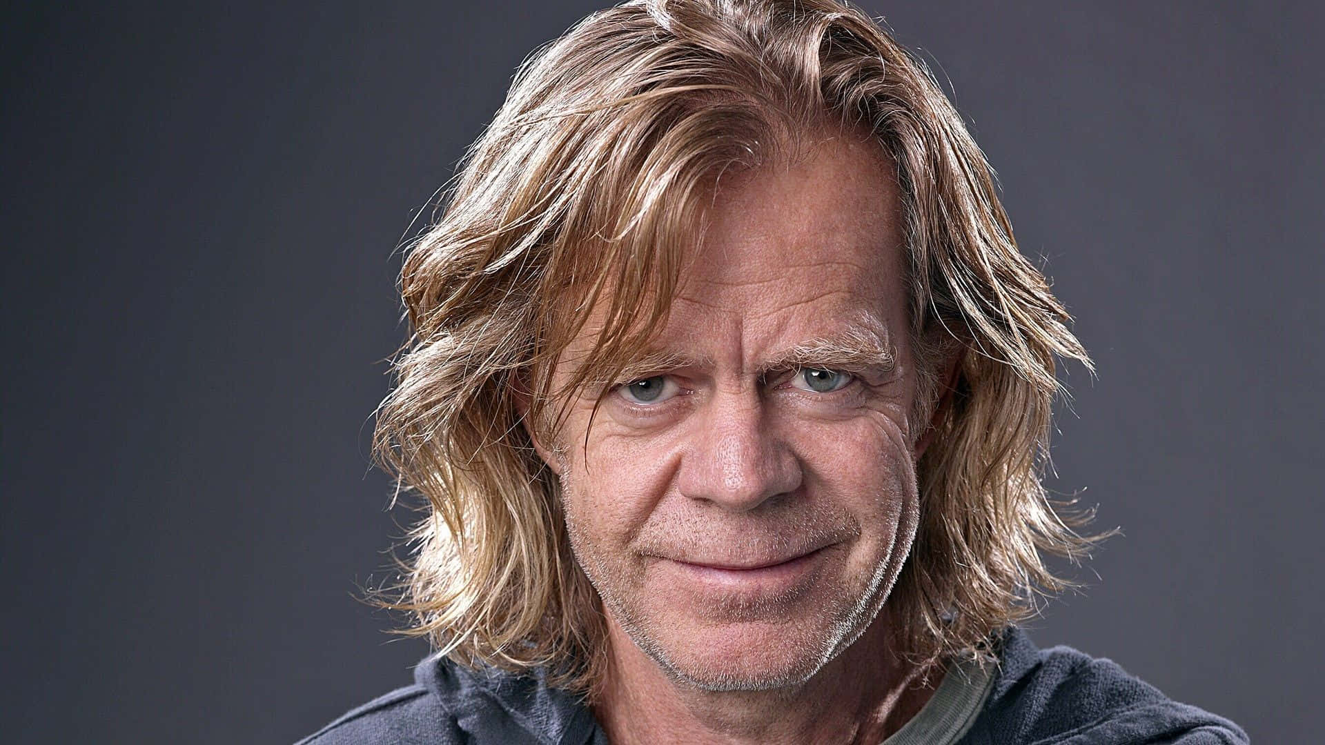 william h. macy 桌布 壁紙 — 一個中年男人的特寫肖像，他有波浪形金髮和微妙微笑，背景是深灰色。