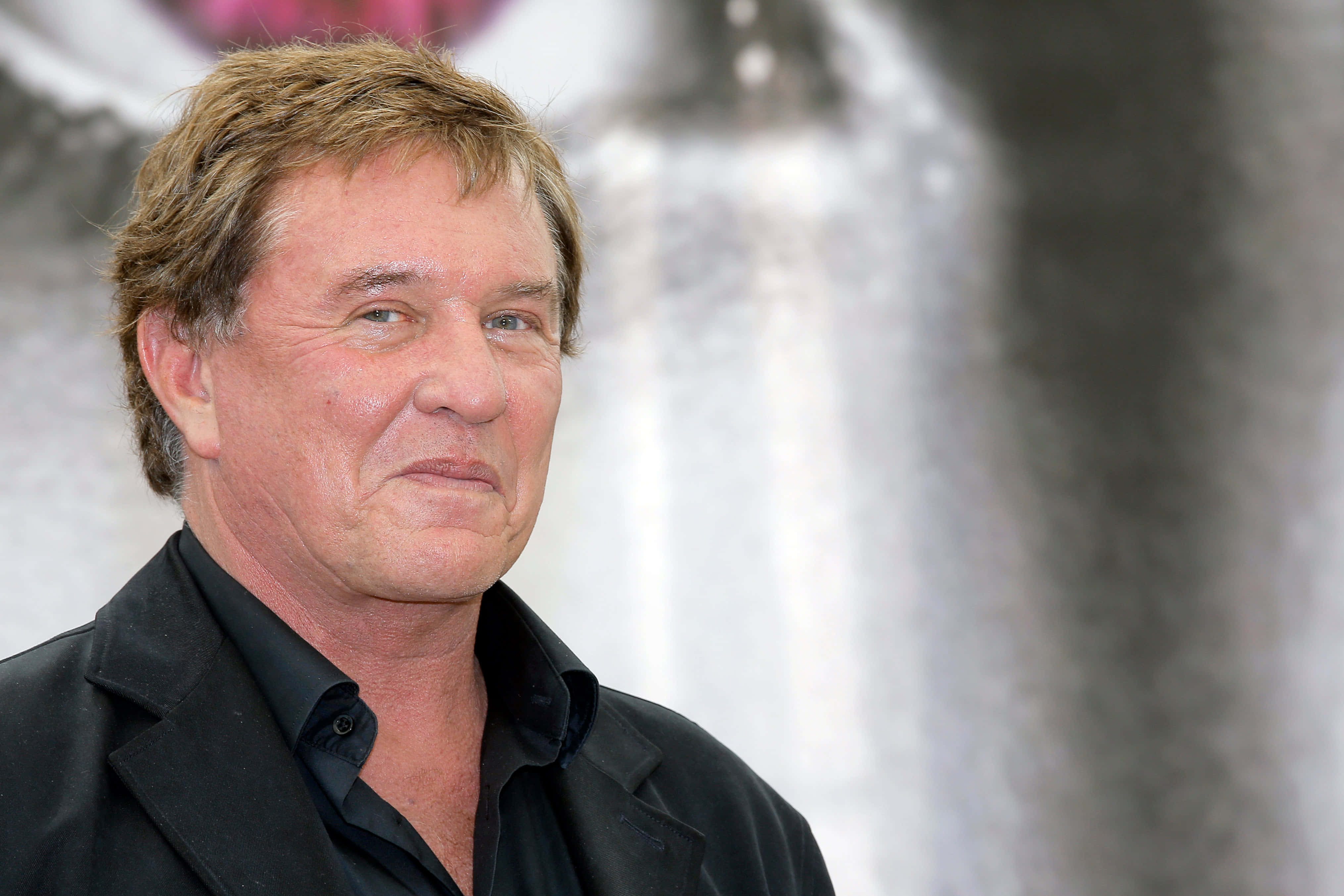 Tom Berenger 桌布 壁紙 — 一個中年男子嘅特寫肖像，佢有亂糟糟嘅棕色頭髮，著住黑色外套，背景係模糊嘅抽象背景。