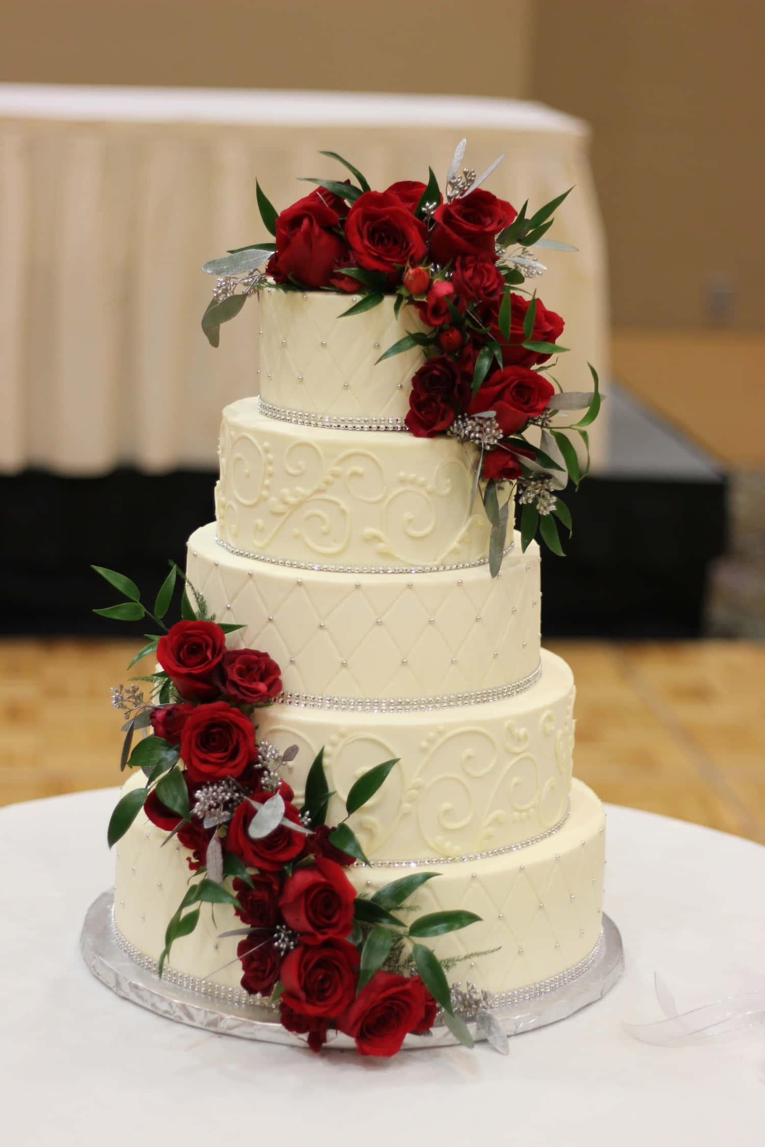 Wedding Cake 桌布 壁紙 — 一個五層象牙色婚禮蛋糕，飾以層疊的紅玫瑰和銀色裝飾，優雅地擺放在白色桌子上。