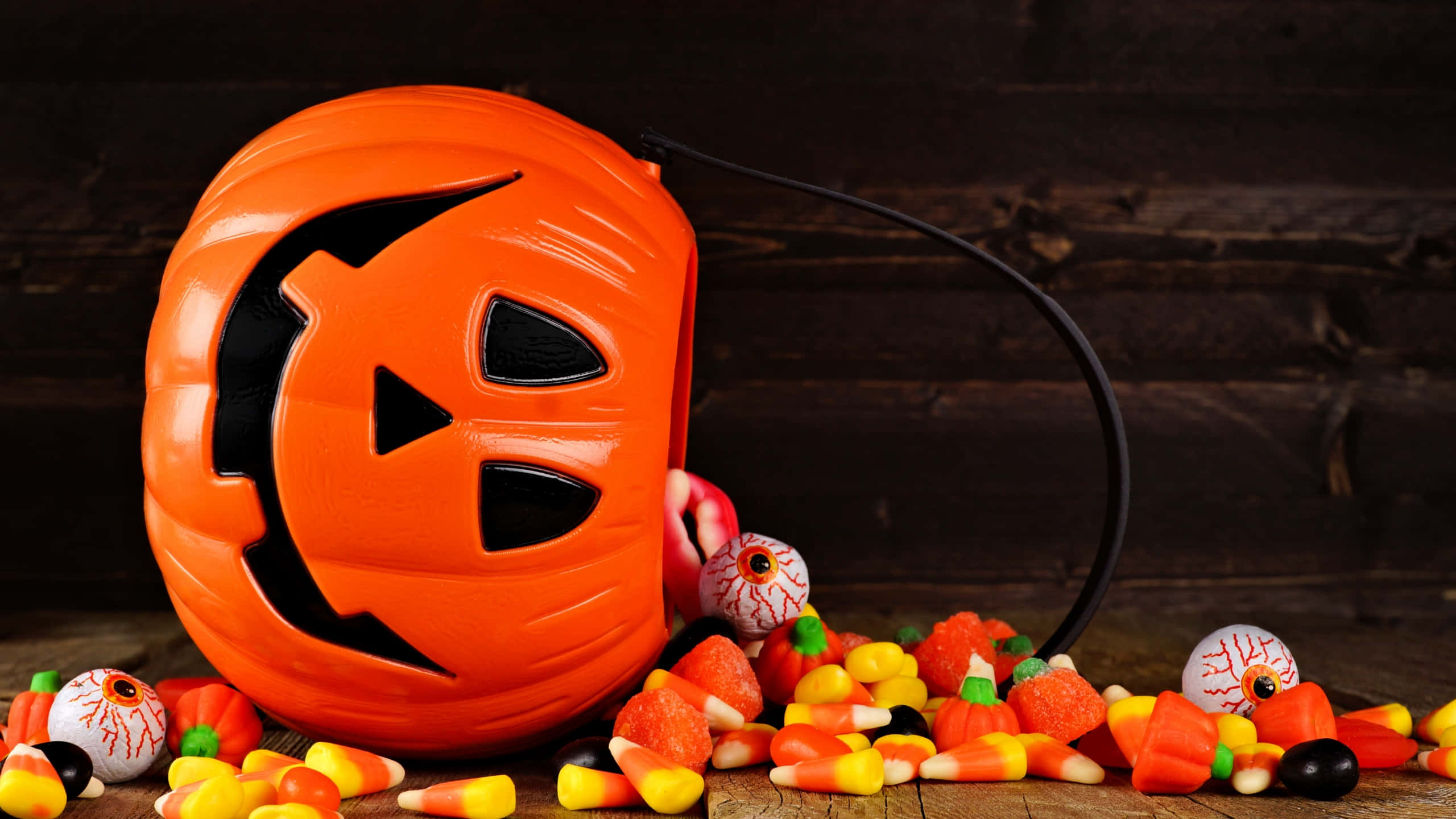 Halloween Treats 桌布 壁紙 — 一個亮橙色南瓜形狀的萬聖節糖果桶，帶有恐怖面孔，放在木質表面上，周圍環繞著玉米糖和眼球糖果。