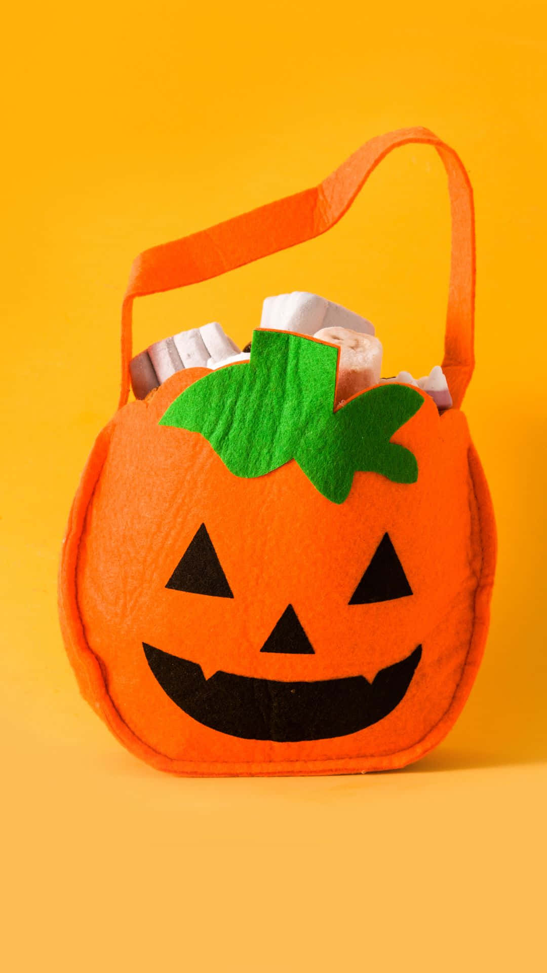 Trick Or Treat Bags 桌布 壁紙 — 一個亮橙色氈製南瓜形狀的萬聖節糖果袋，帶有綠色莖和黑色傑克南瓜燈臉，裝滿包裝好的零食，放在純黃色背景上。