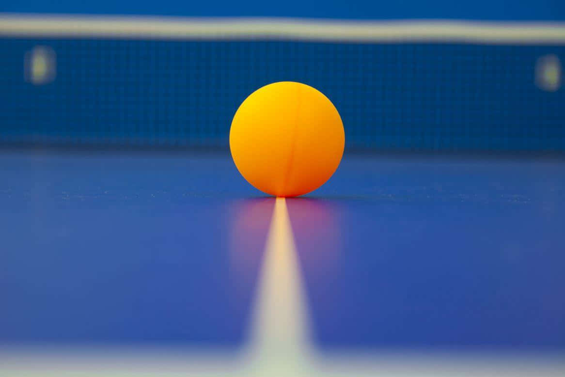 Table Tennis 桌布 壁紙 — 一個亮橙色的乒乓球放在藍色桌子上，後面有白色線條和網，背景柔和模糊。