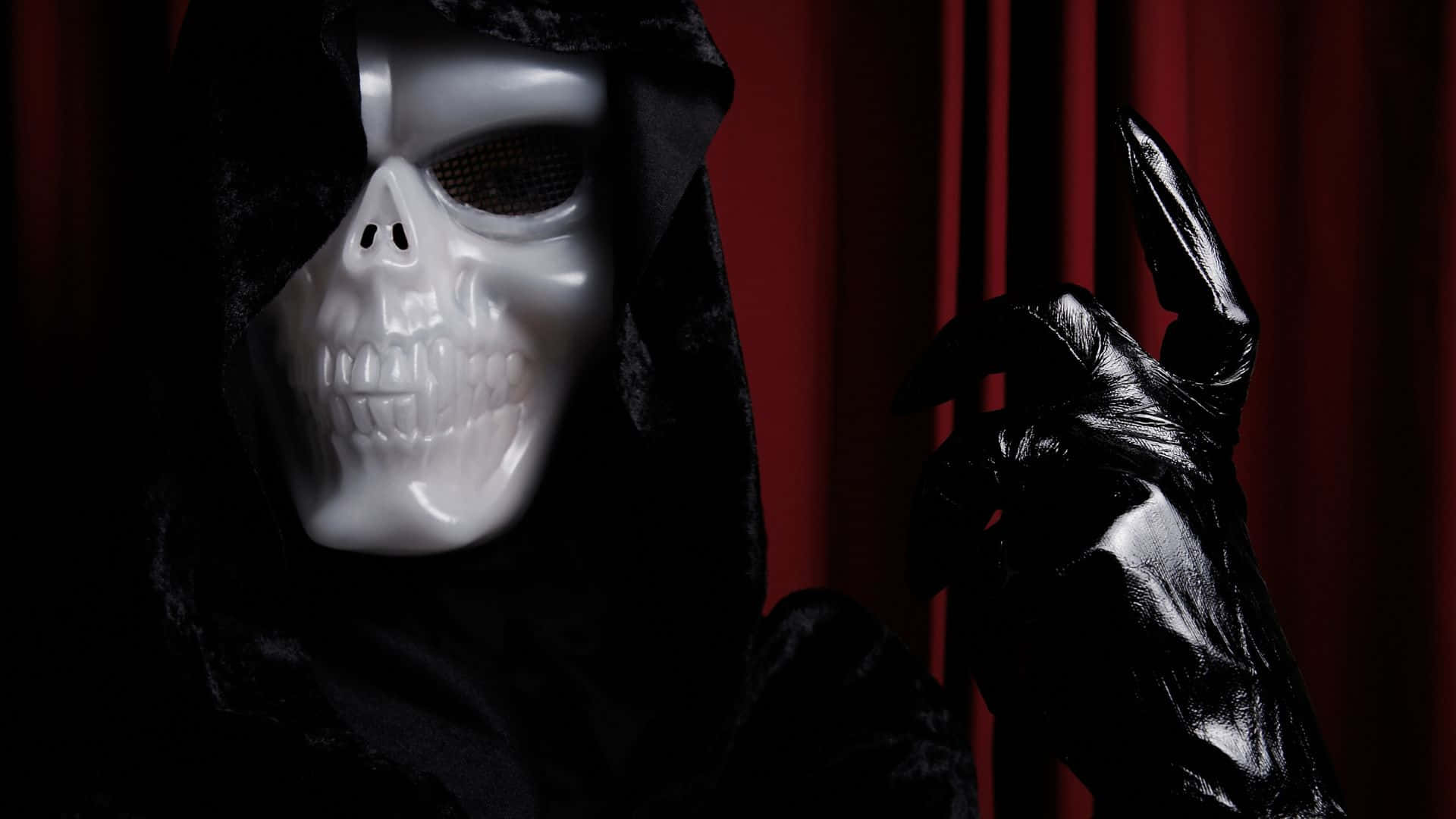 Horror Costumes 桌布 壁紙 — 一個亮面白色骷髏面具，帶有黑色網眼眼睛，一個黑色連帽身影在深紅色天鵝絨窗簾前舉起戴手套的手。