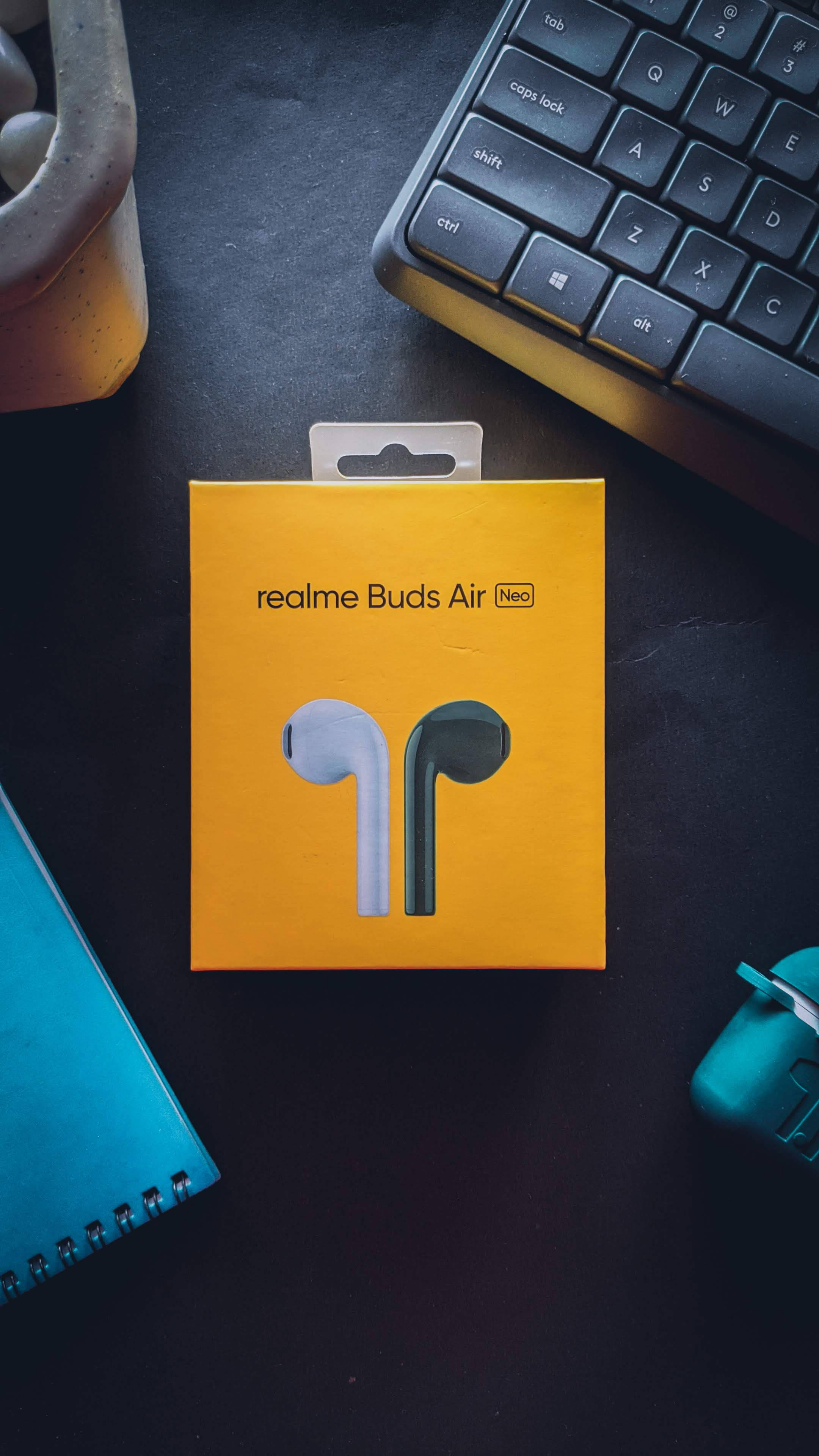 Realme Buds Air 壁紙 — 一個亮黃色的 realme Buds Air Neo 盒子放在深色書桌中央，旁邊有鍵盤、筆記本和青綠色滑鼠。