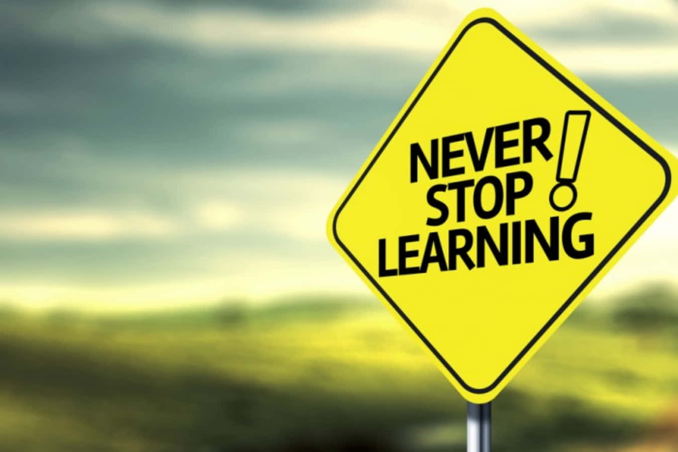 學習 桌布 壁紙 — 一個亮黃色菱形路牌，上面有粗體黑色文字寫著「NEVER STOP LEARNING!」，在陰天背景下，以模糊的自然景觀為襯托。