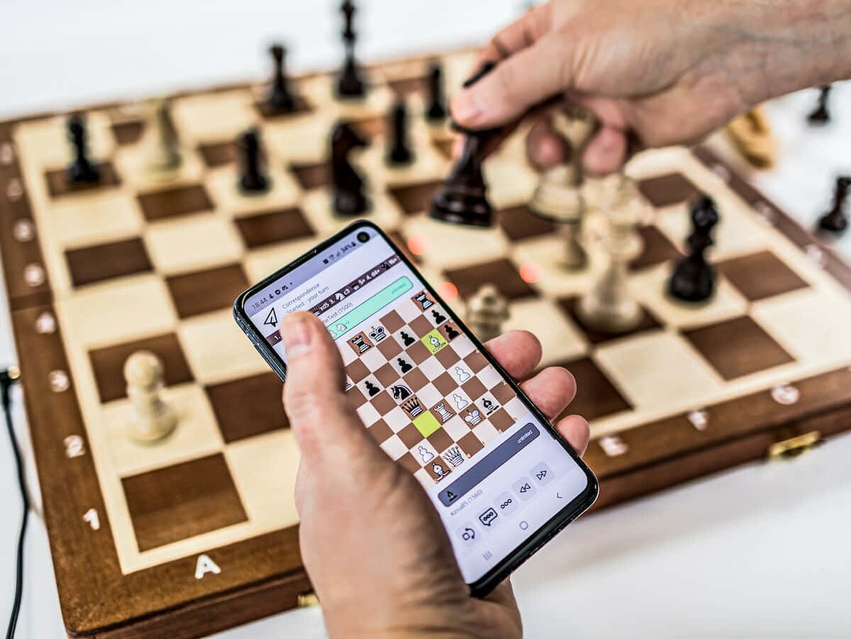Chessboard 桌布 壁紙 — 一個人下棋，融合實體棋盤和智能手機應用程式，專注的手放在木棋盤和發光屏幕上。