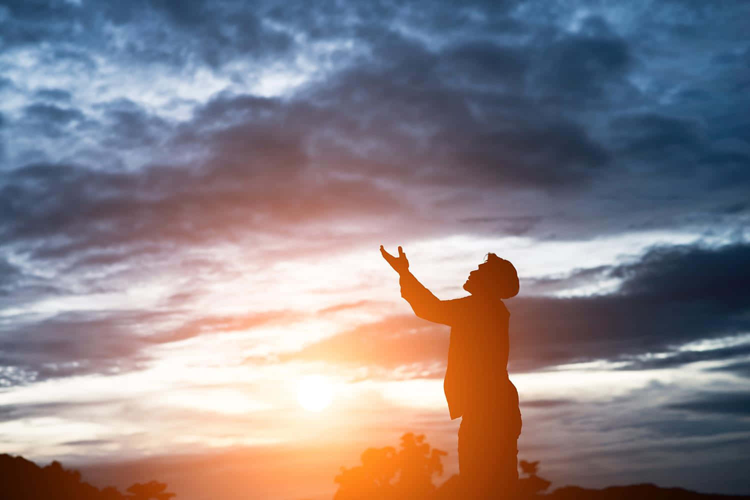 Praying 桌布 壁紙 — 一個人以剪影站立，雙臂舉向充滿橙色和藍色雲彩的戲劇性日落天空。