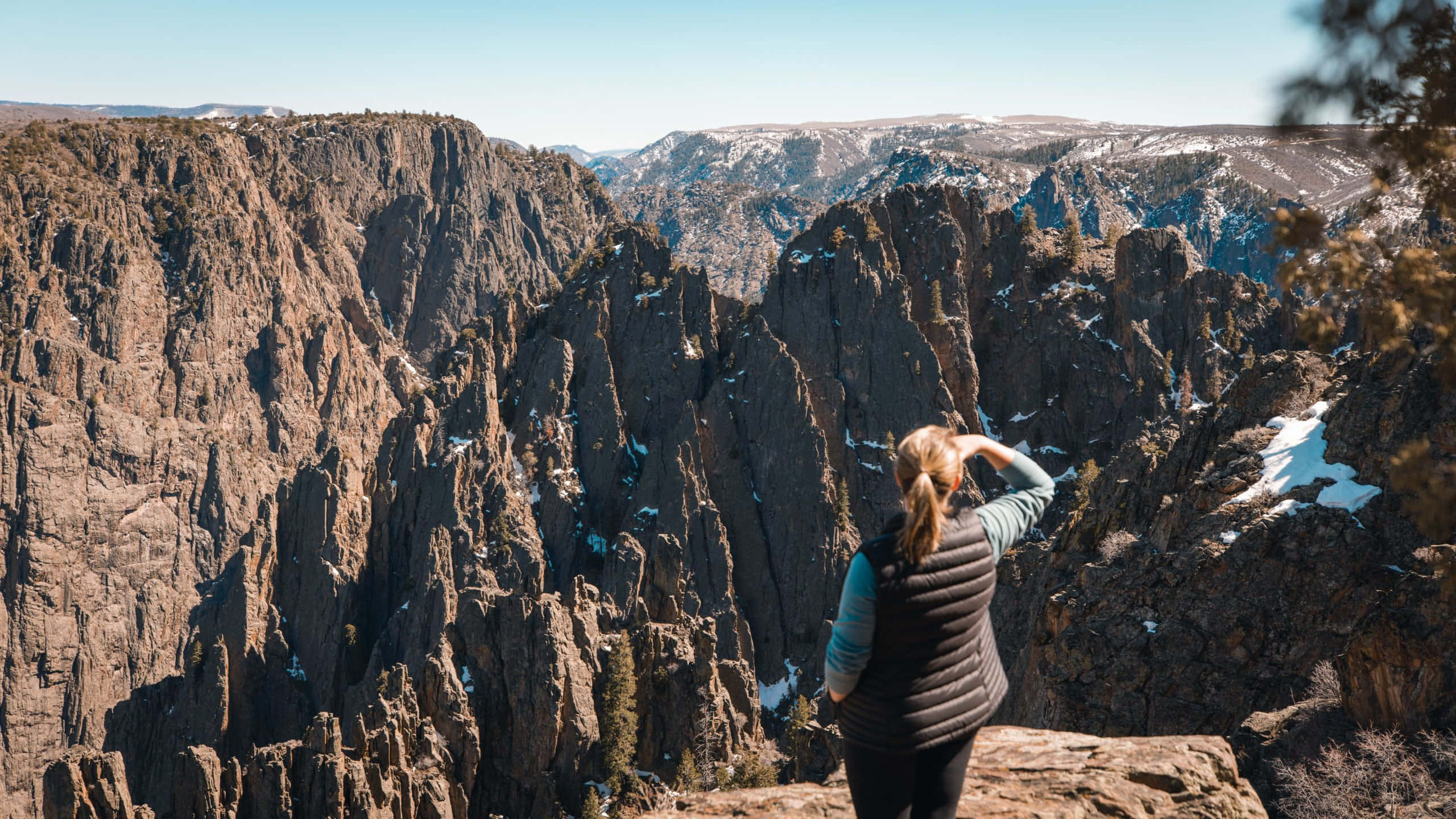 Black Canyon 桌布 壁紙 — 一個人企喺岩石懸崖邊，俯瞰廣闊崎嶇嘅峽谷，白雪覆蓋嘅山峰喺晴朗藍天下。