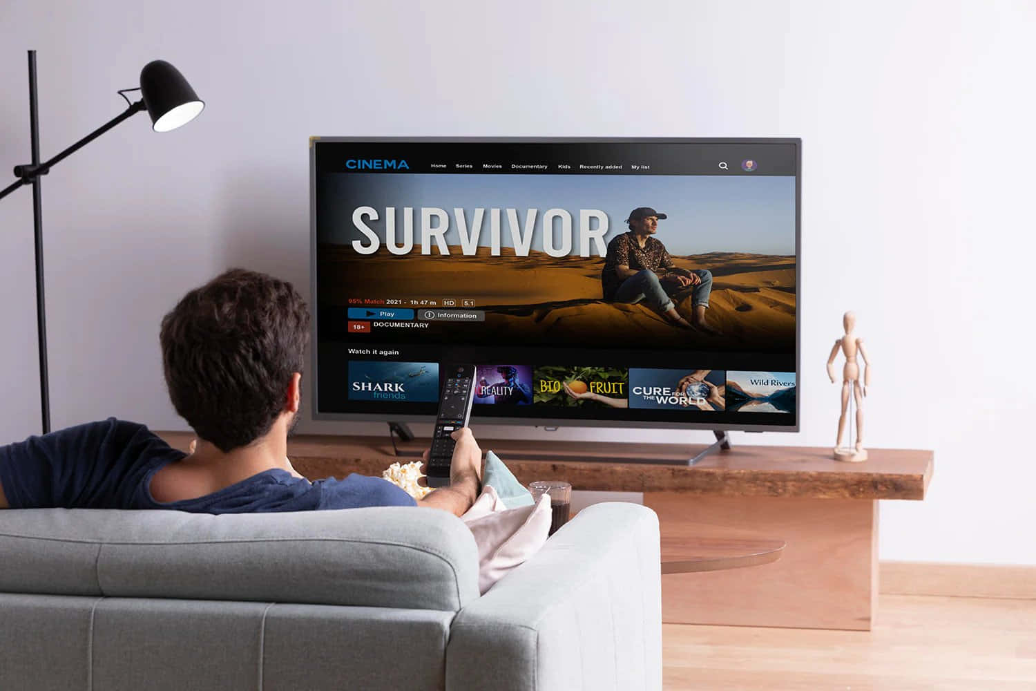 tv 桌布 壁紙 — 一個人喺梳化上放鬆，睇緊電視播放緊嘅節目《Survivor》，有沙漠場景同一個木製人體模型喺電視旁邊。