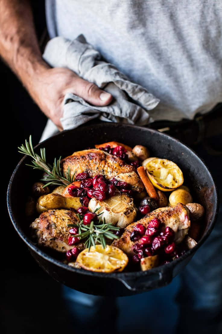 Minimalist Food 桌布 壁紙 — 一個人喺溫暖嘅金色燈光下，揸住一個鑄鐵煎鍋，上面有烤雞、小紅莓、迷迭香同柑橘。
